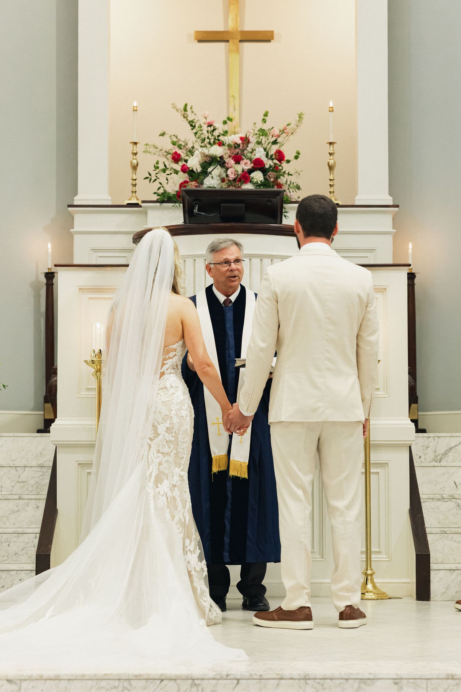Bride and groom hold hands with officiant at church altar beneath golden cross during wedding ceremony.