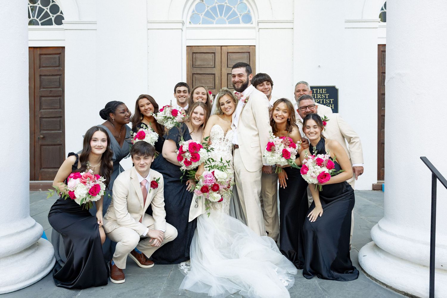 Wedding party posing outside church with bride in white gown and bridesmaids holding pink bouquets.