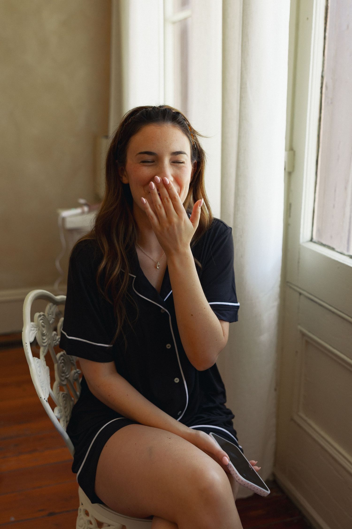 Young woman in black button-up pajama dress laughing with hand over mouth, seated on ornate chair by window.