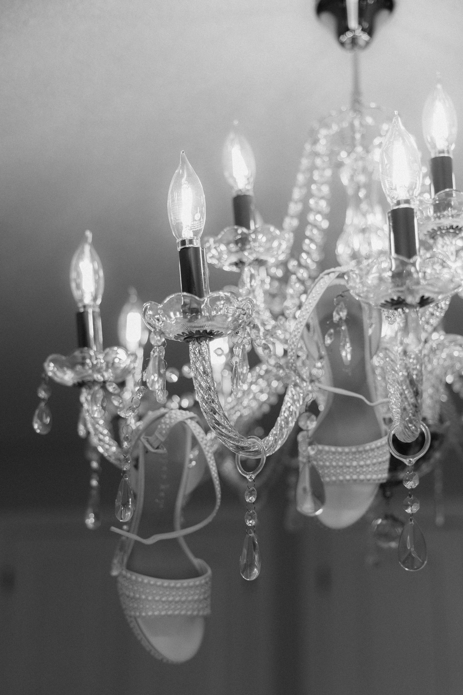 Black and white photo of elegant crystal chandelier with lit candles and a strappy heeled sandal hanging from it.