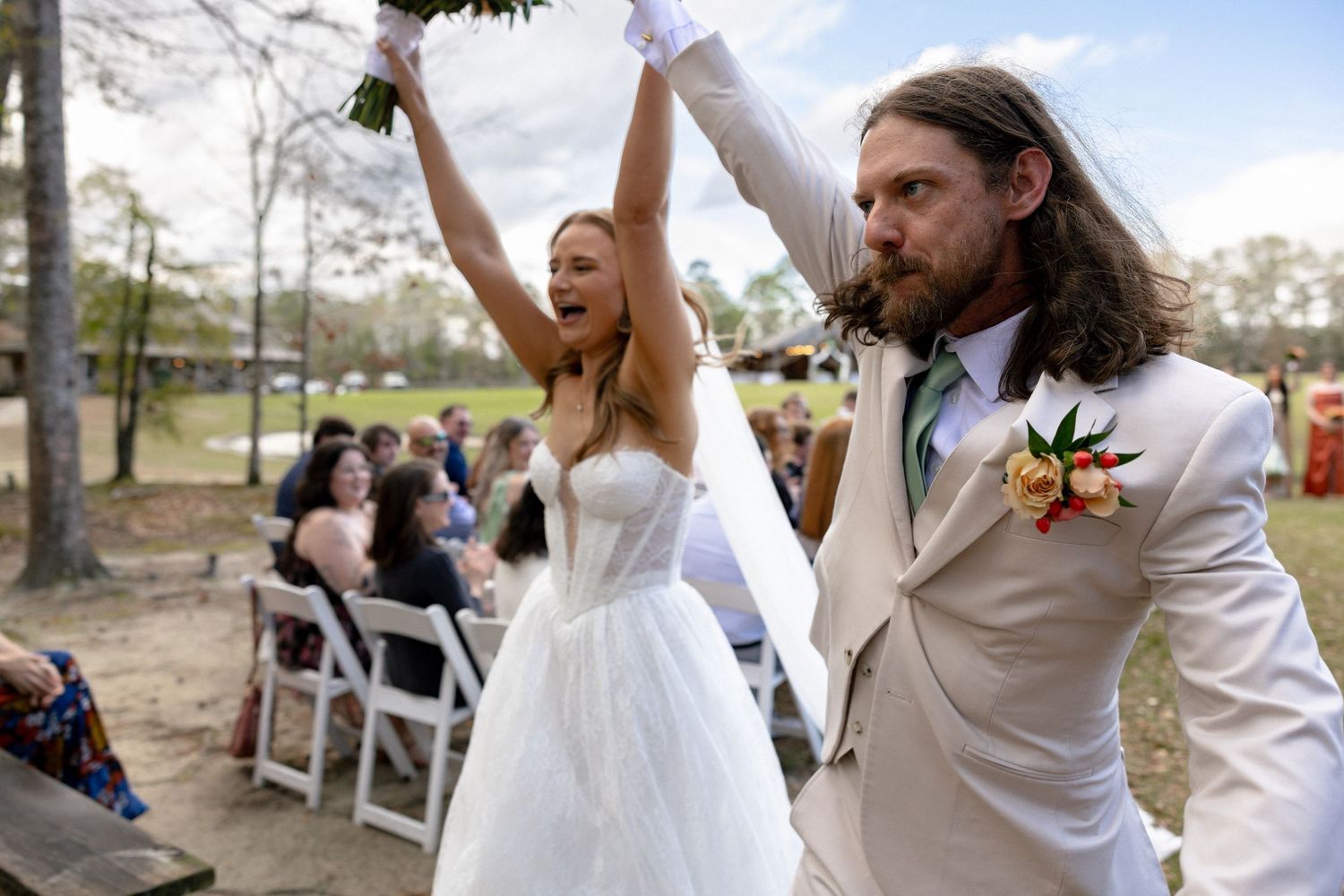 Excited newlyweds celebrate walking down aisle at outdoor wedding, bride in white gown, groom in cream suit.