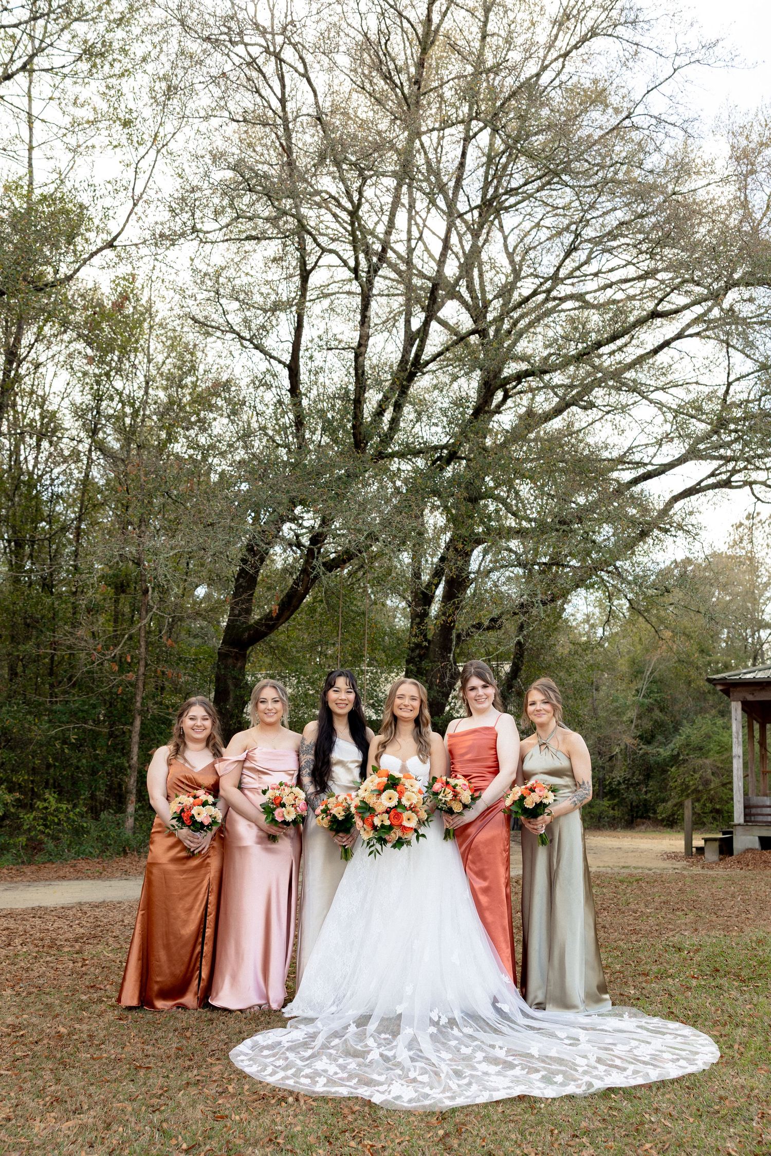 Bride in lace gown stands with five bridesmaids in mismatched earth-tone satin dresses holding floral bouquets.