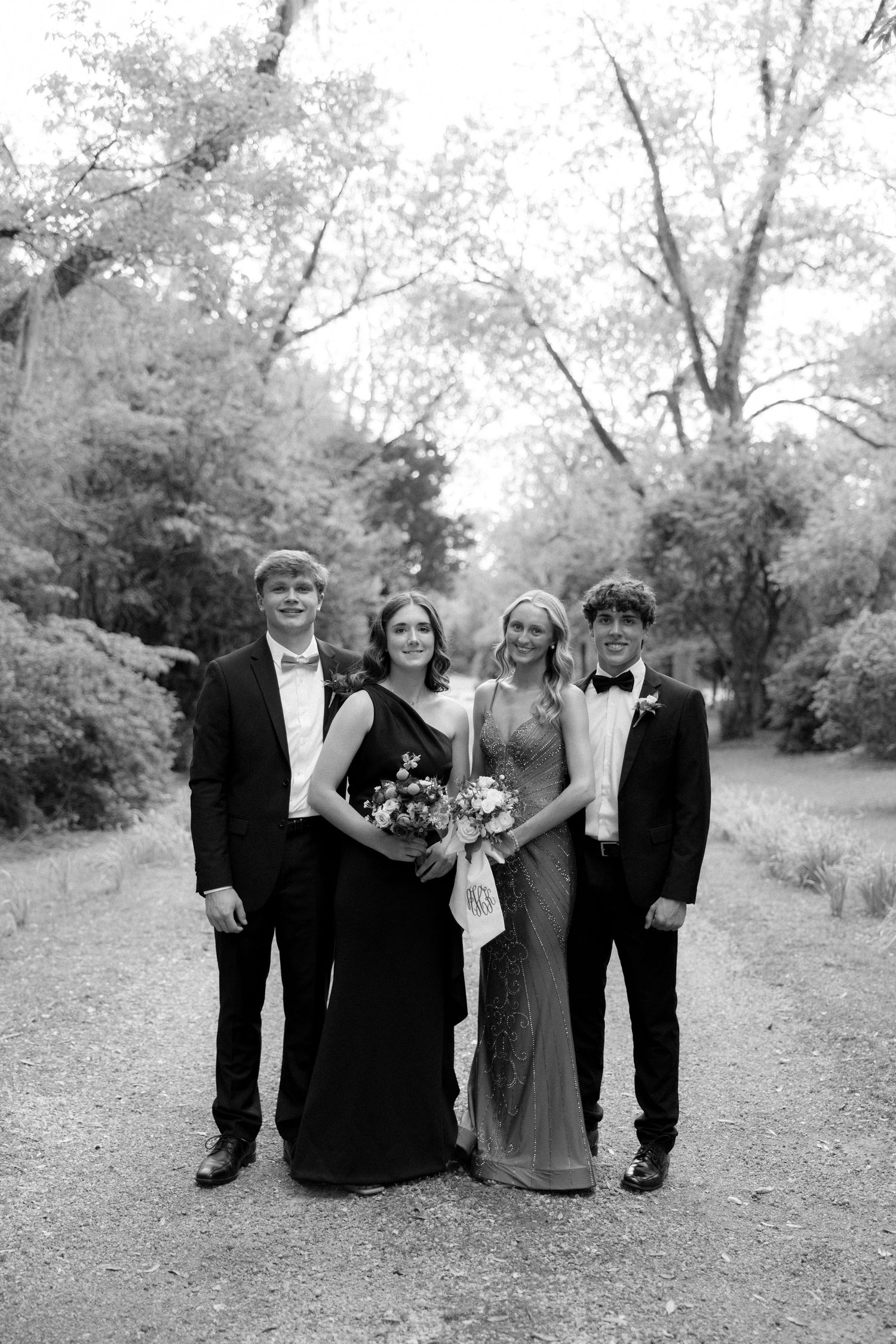 Black and white photo of two prom couples posing together on a tree-lined path.