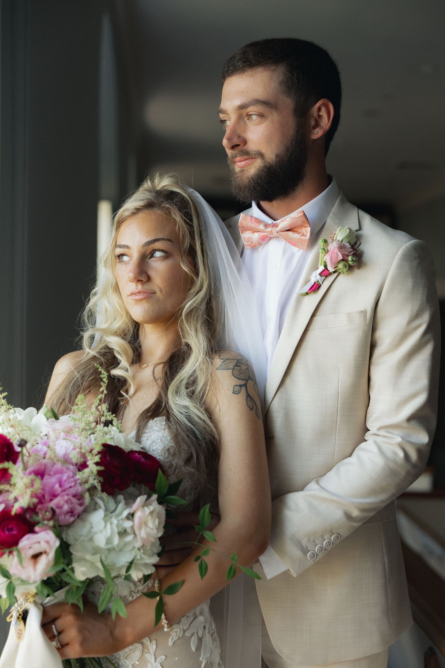 Bride holding colorful bouquet stands with bearded groom in beige suit and pink bow tie near window.