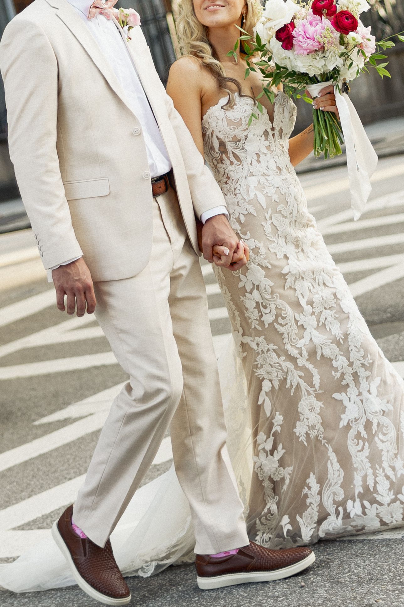 Bride and groom walking hand in hand, she in lace gown holding bouquet, he in cream suit with brown loafers.