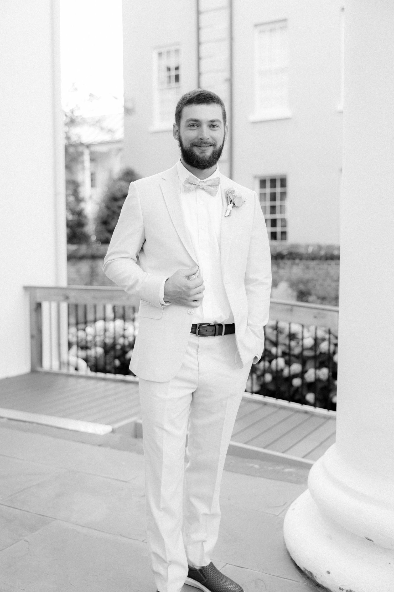 Bearded groom in cream suit and bow tie stands confidently on outdoor venue porch, black and white.