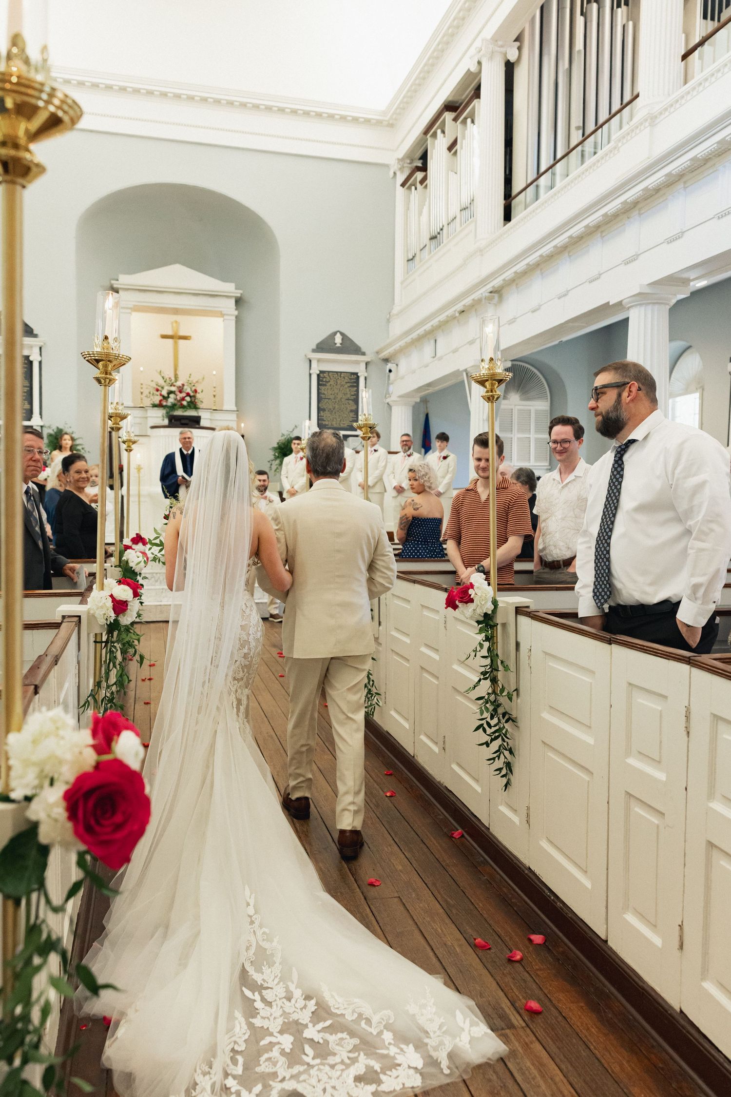 Bride in lace gown with long veil walks down decorated church aisle with escort during wedding ceremony.