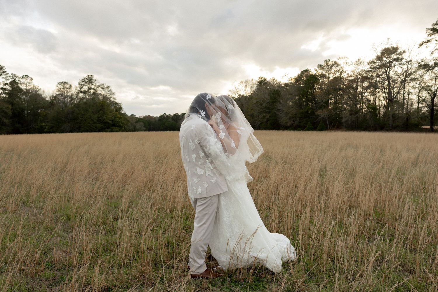 Bride and groom share a passionate kiss under a veil in a golden meadow at dusk.