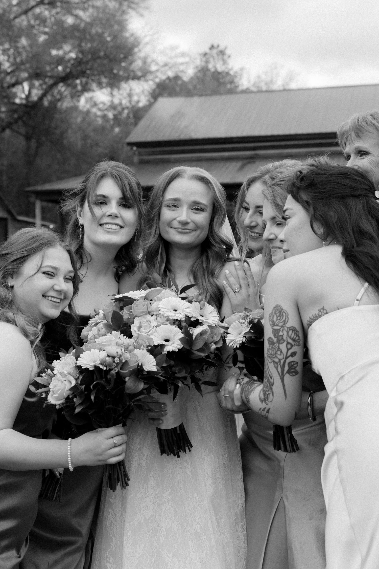 Black and white photo of smiling bride surrounded by bridesmaids holding bouquets outside a rustic wedding venue.