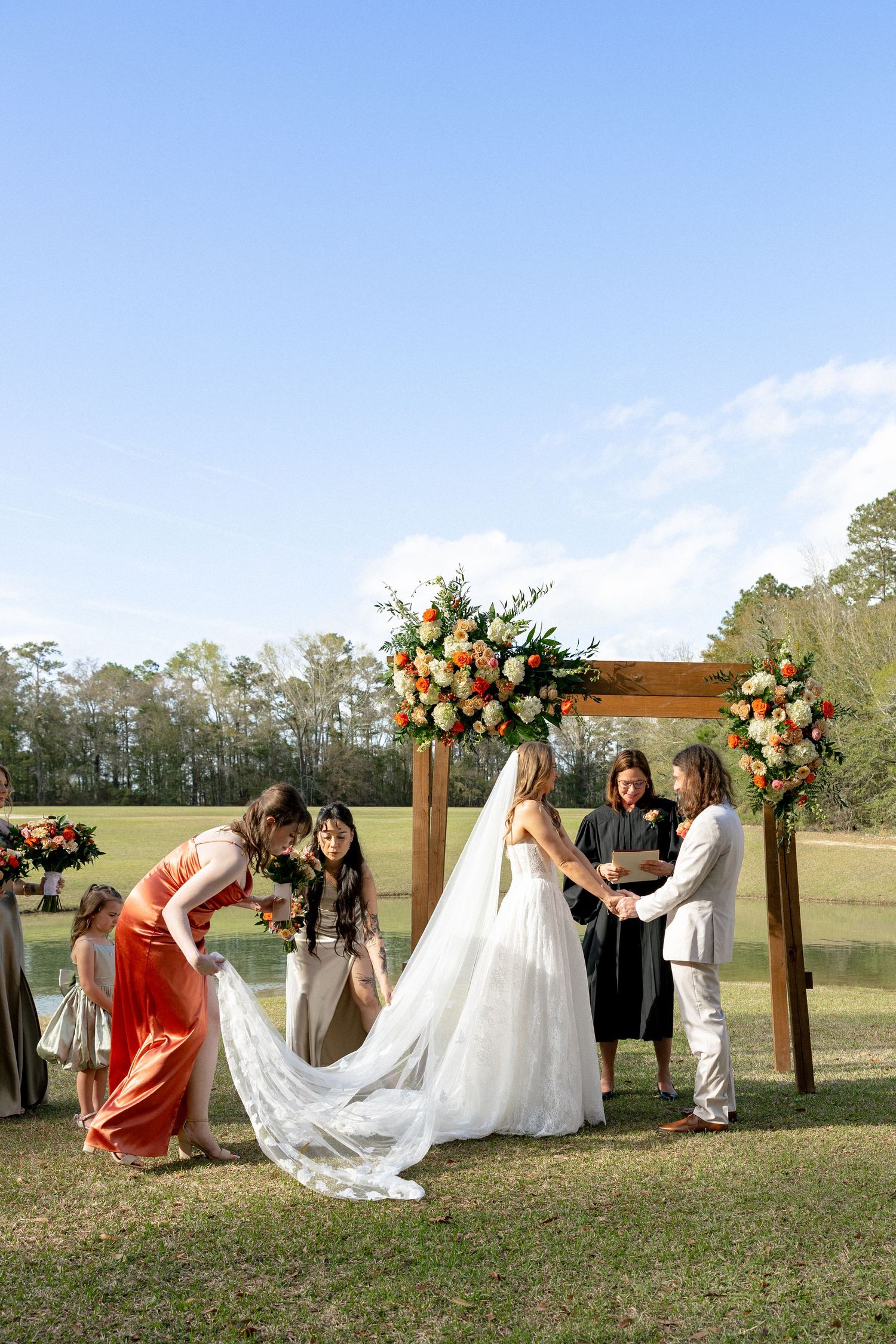 Bridesmaids adjusting bride's train at outdoor wedding ceremony beneath wooden arch with floral arrangements.