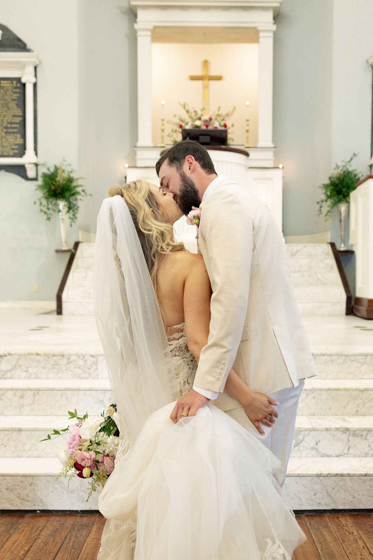 Bride and groom share a romantic kiss at the altar of a church, bride holding a pink and white bouquet.