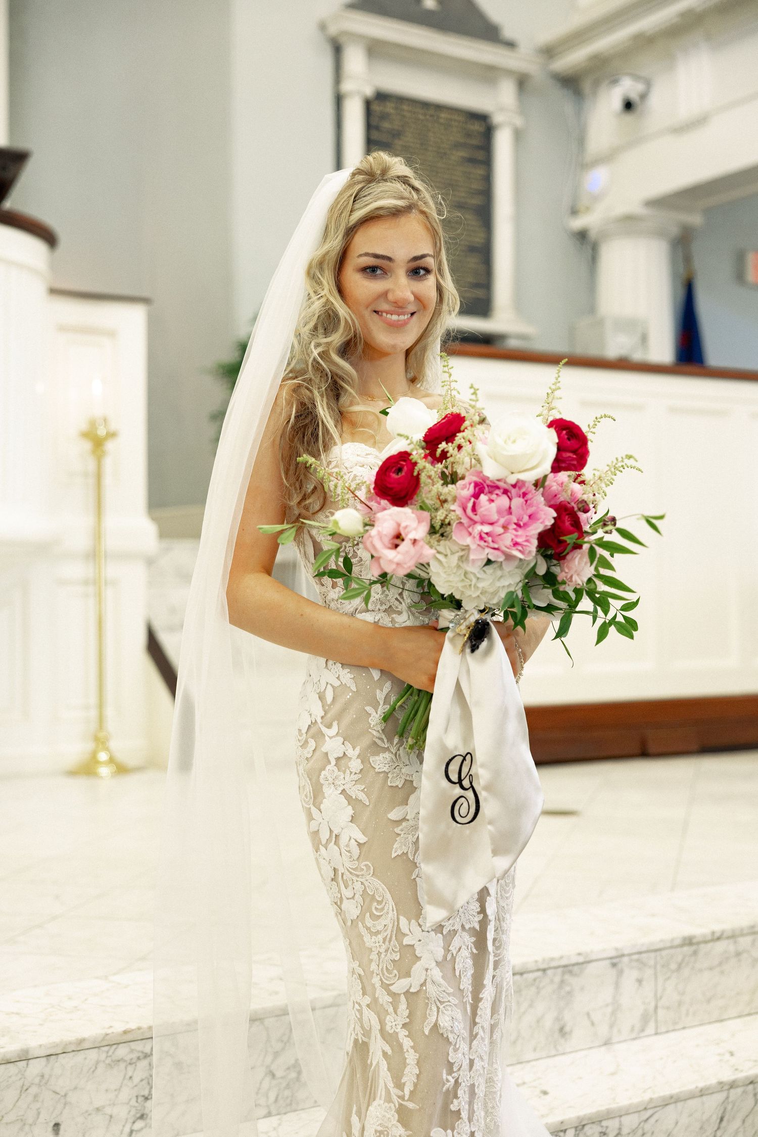 Smiling bride in lace mermaid gown holds pink and red bouquet with monogram ribbon in white church.