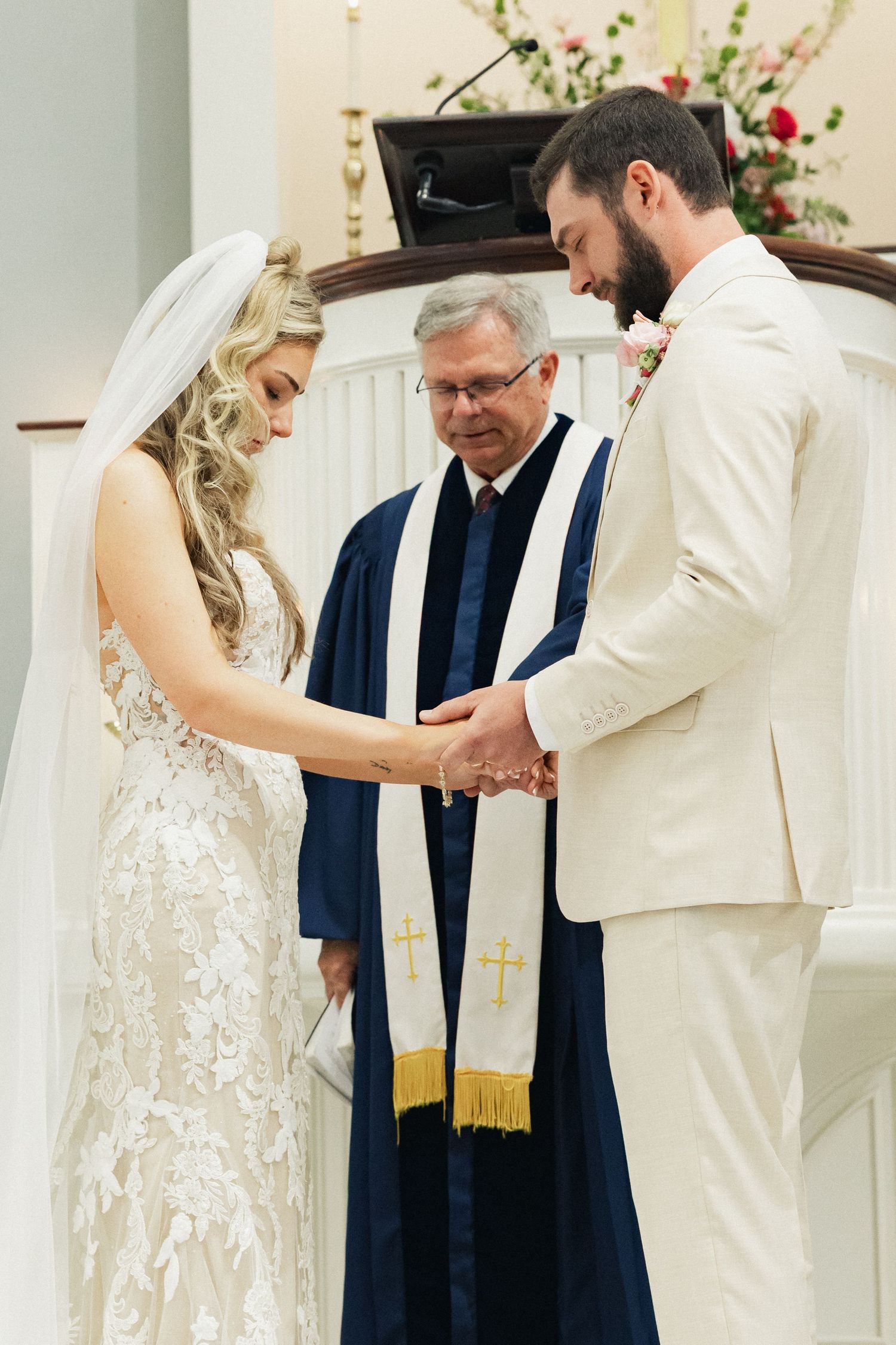 Bride and groom bow their heads in prayer while holding hands before a robed officiant at the altar.