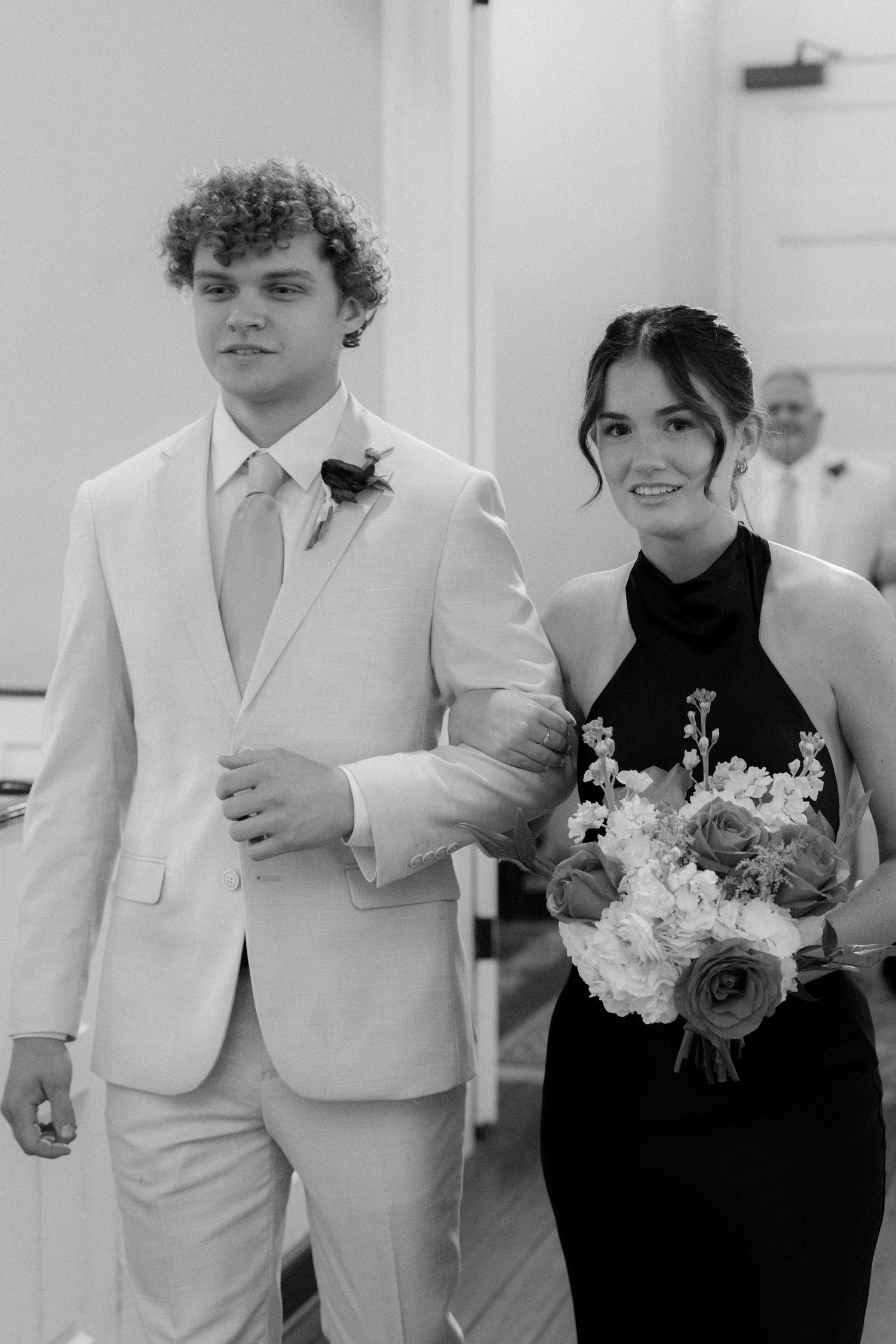 Young groomsman in light suit escorts bridesmaid in black dress holding bouquet down wedding aisle, black and white photo.