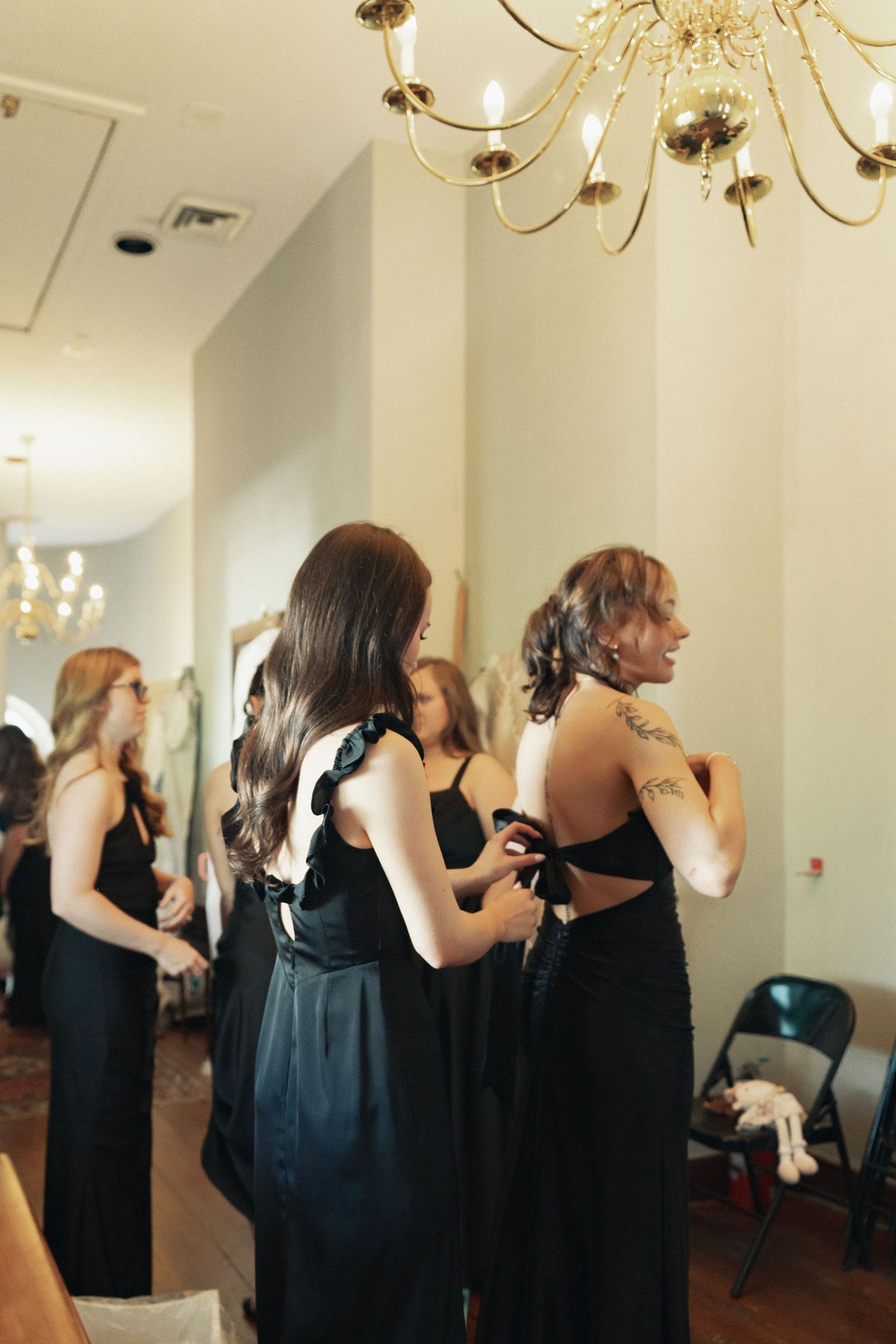 Bridesmaids in elegant black dresses help each other get ready under a gold chandelier in a bright room.