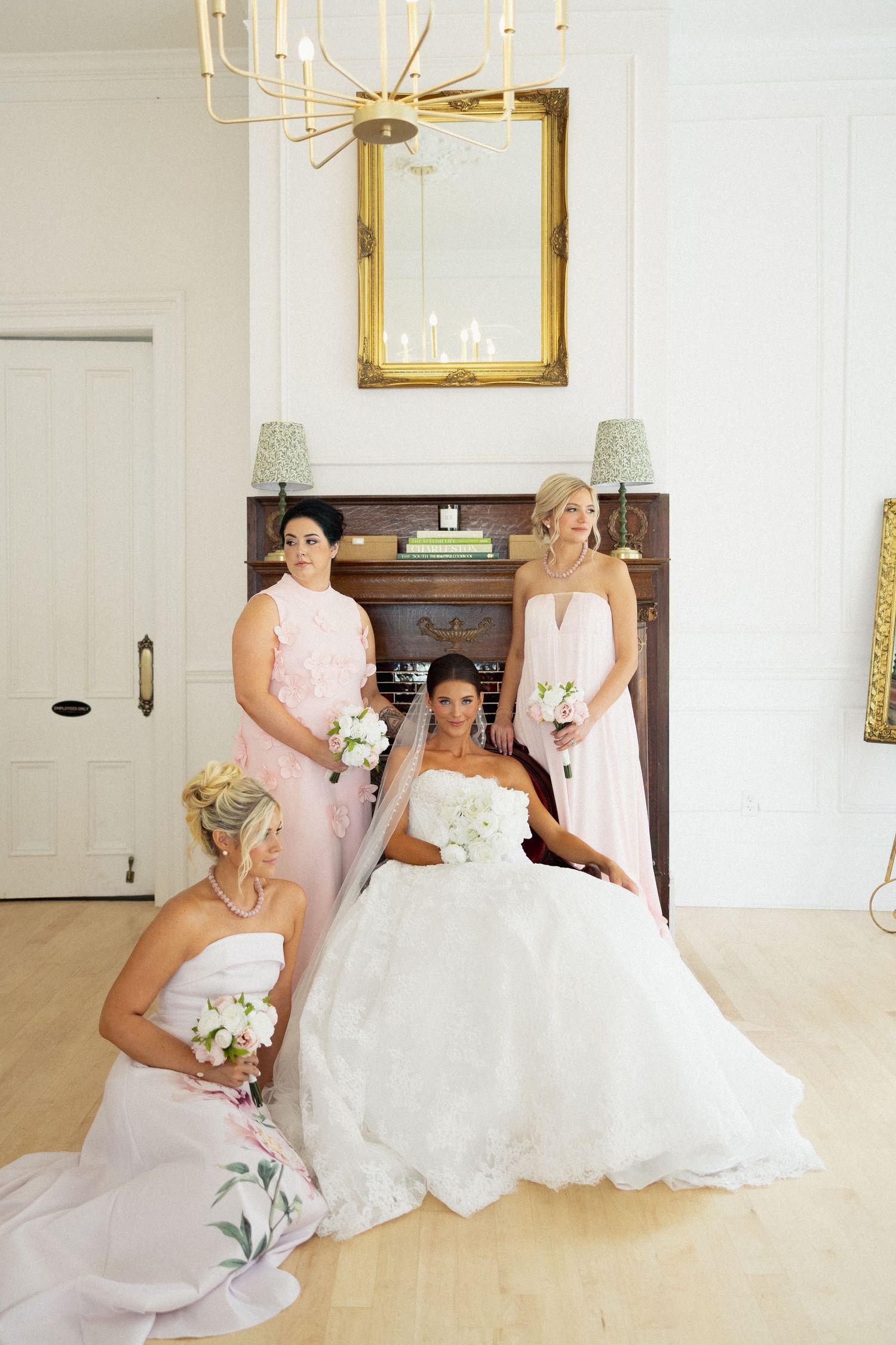 Bride in white ball gown poses with three bridesmaids in pink dresses holding bouquets in elegant white room.