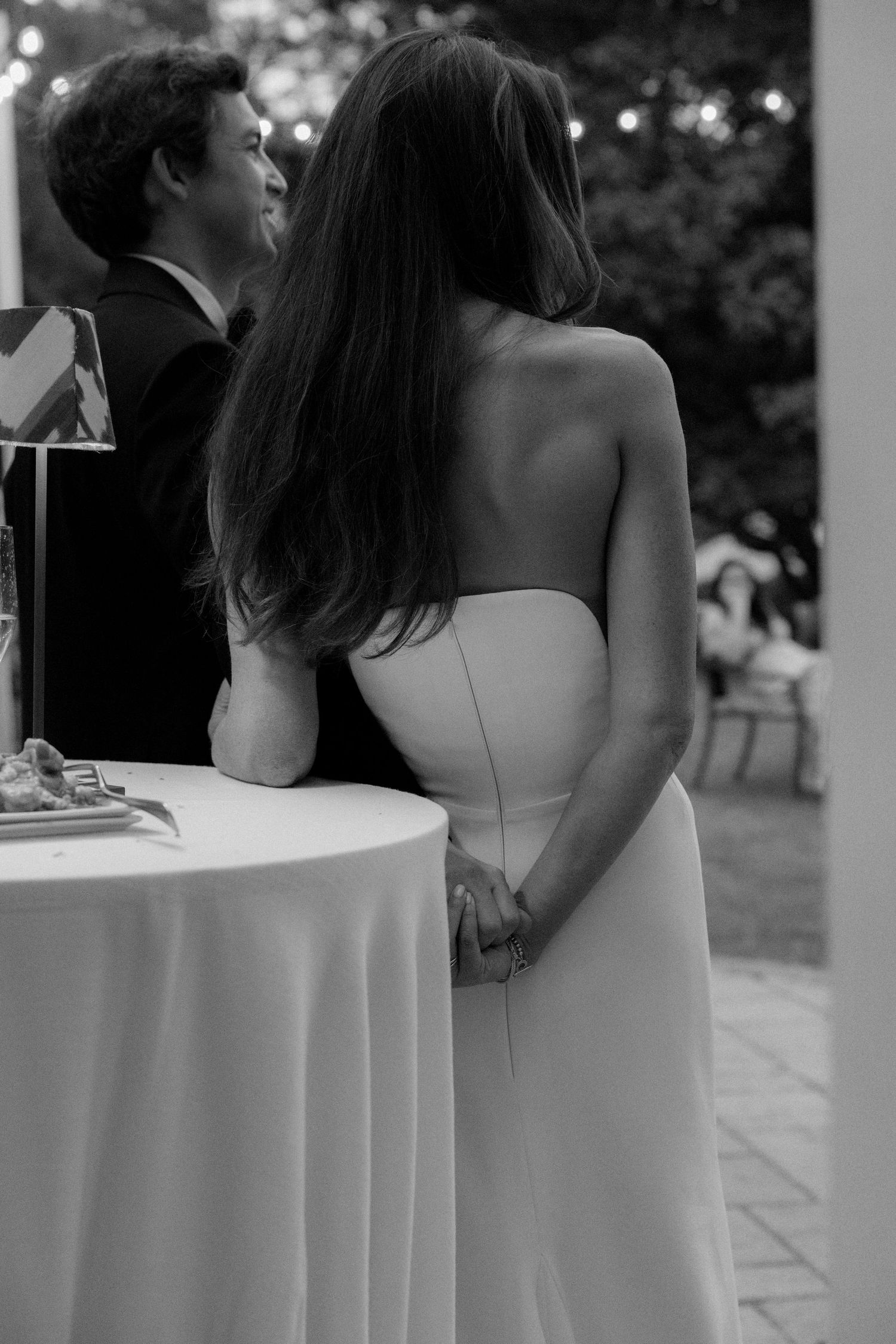 Black and white photo of a bride in strapless gown holding hands with groom at wedding reception table.