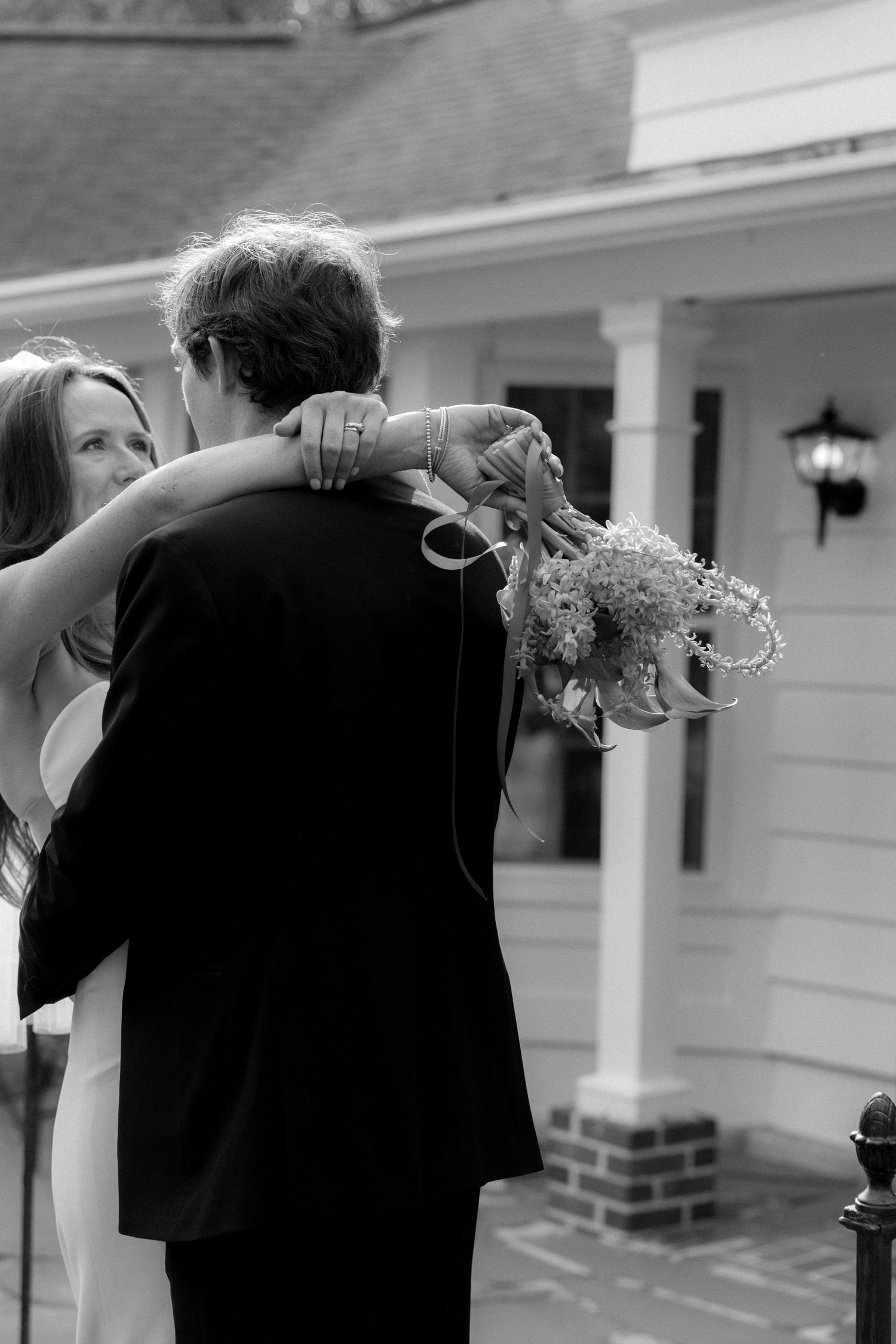 Black and white photo of bride and groom embracing outdoors, holding a bouquet, in front of a classic white home.