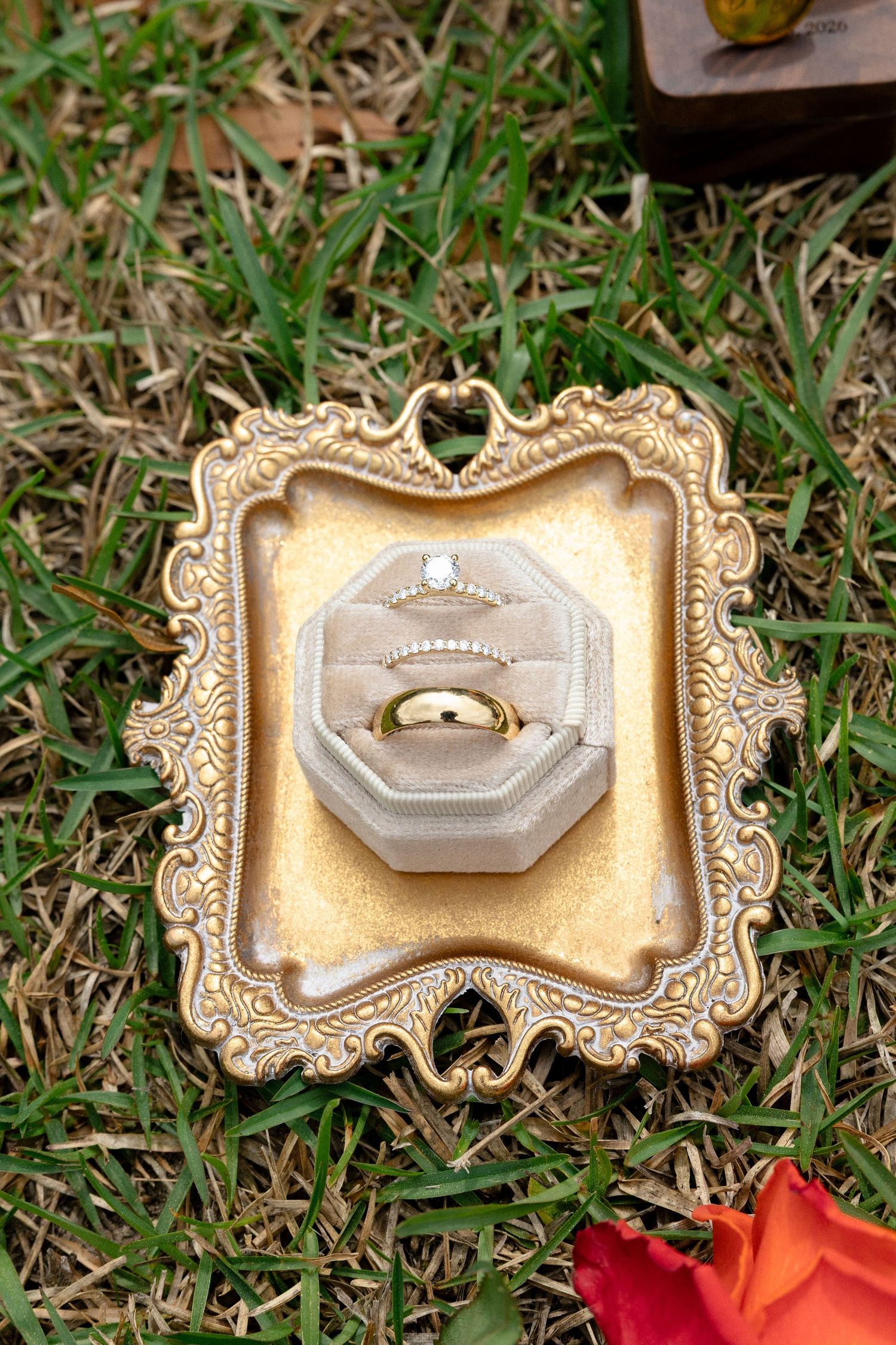 Gold wedding band and diamond engagement ring in velvet box displayed on ornate antique gold tray on grass.