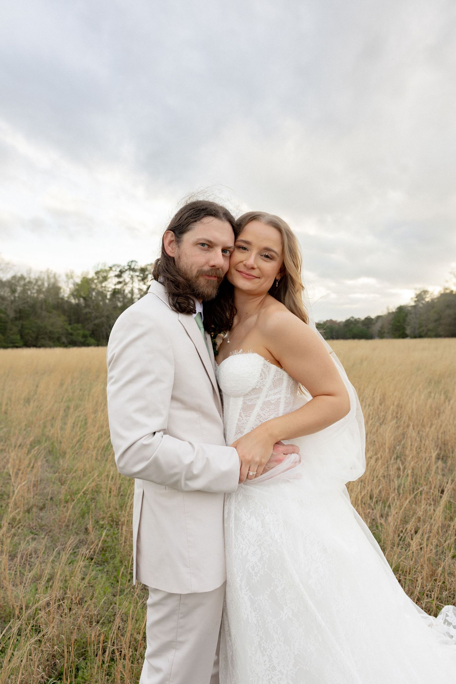 Bride and groom pose together in golden grass field, bride in strapless lace wedding gown.