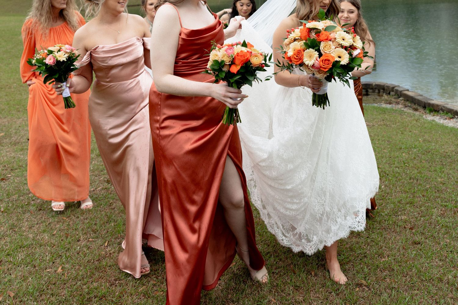 Bridesmaids in rust and blush satin gowns walk alongside bride in white lace dress near a lakeside venue.