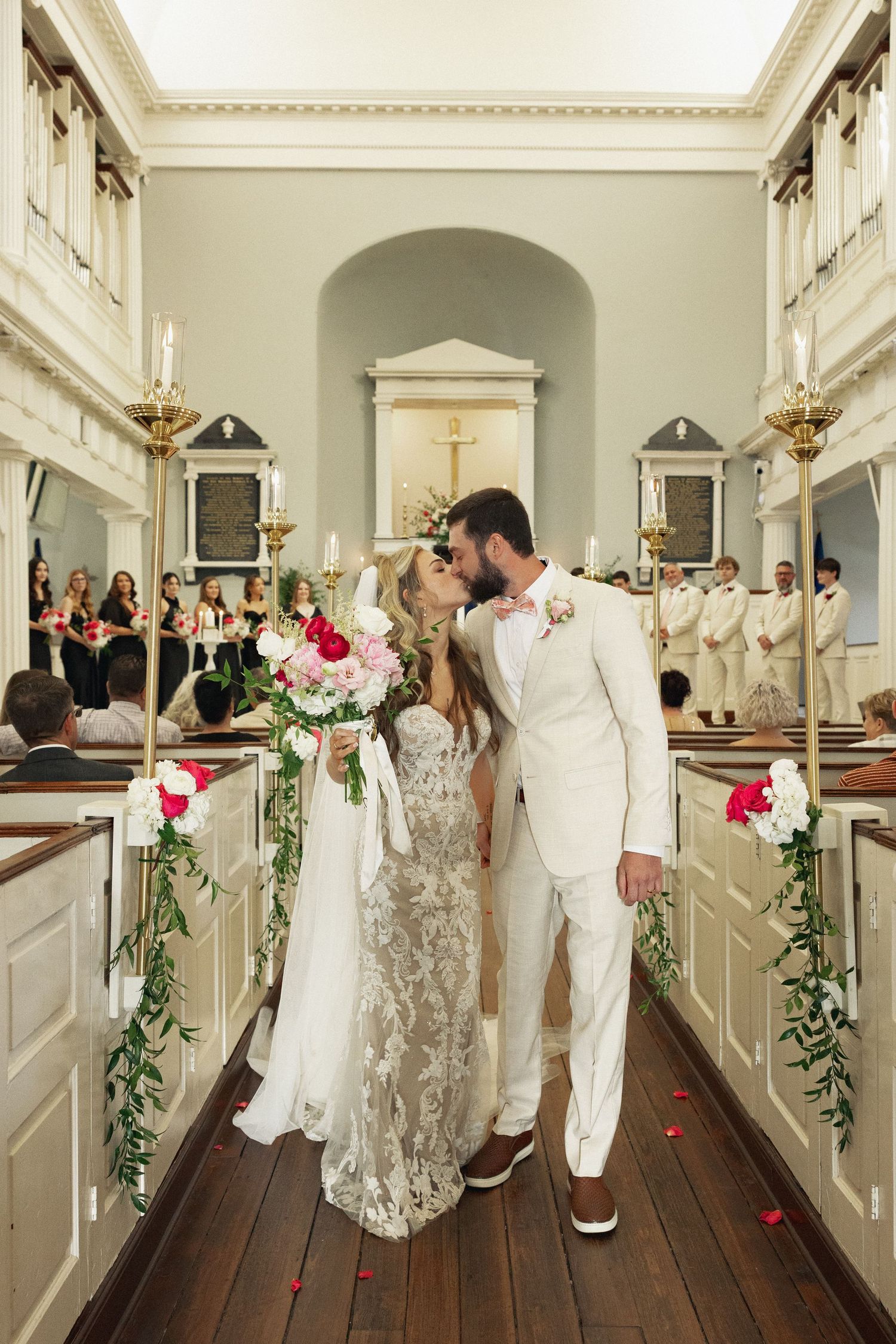 Bride and groom share first kiss in elegant white church aisle with rose petal decor.