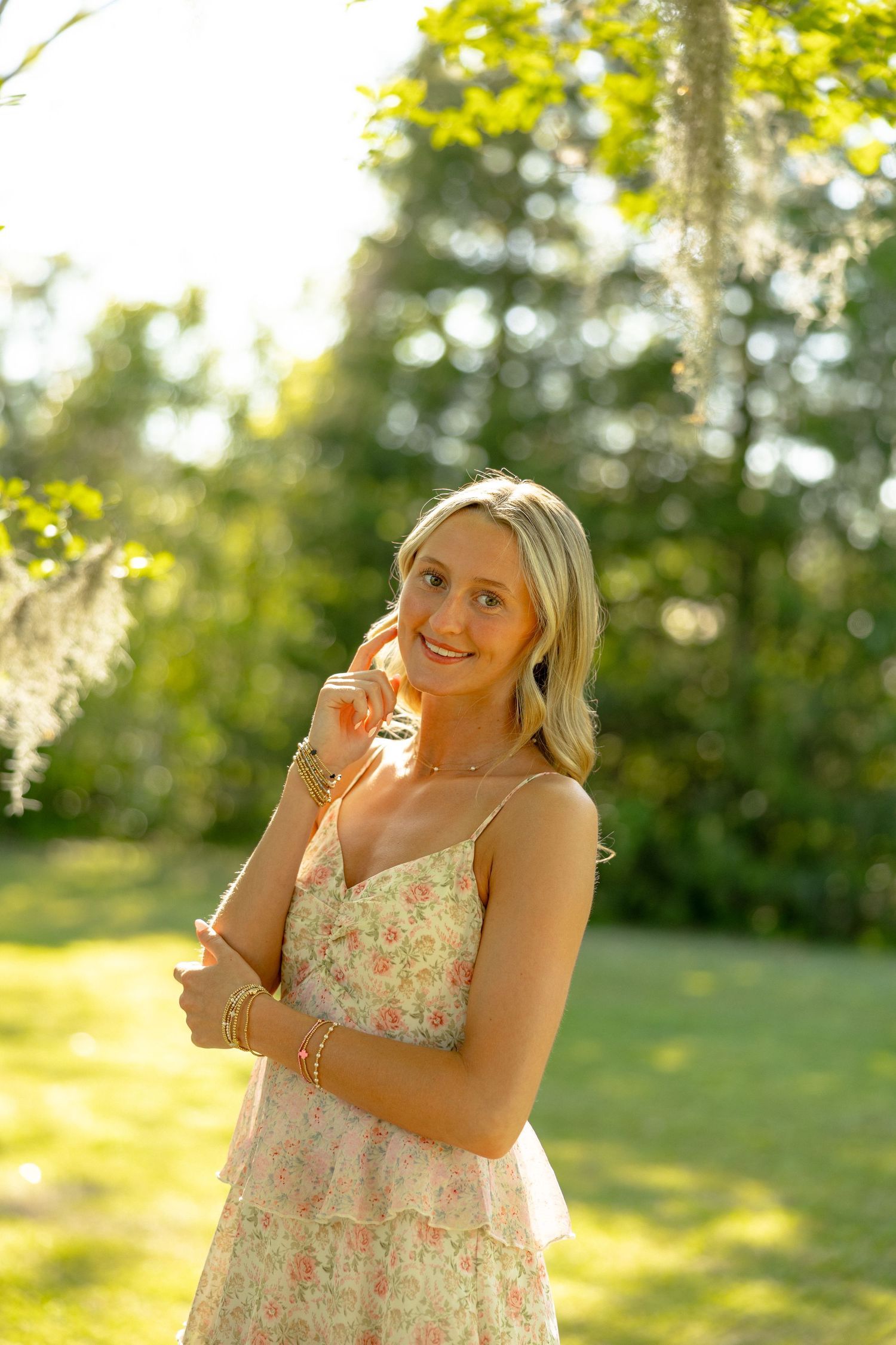 Smiling blonde woman in a floral sundress posing outdoors in a sunlit green garden setting.