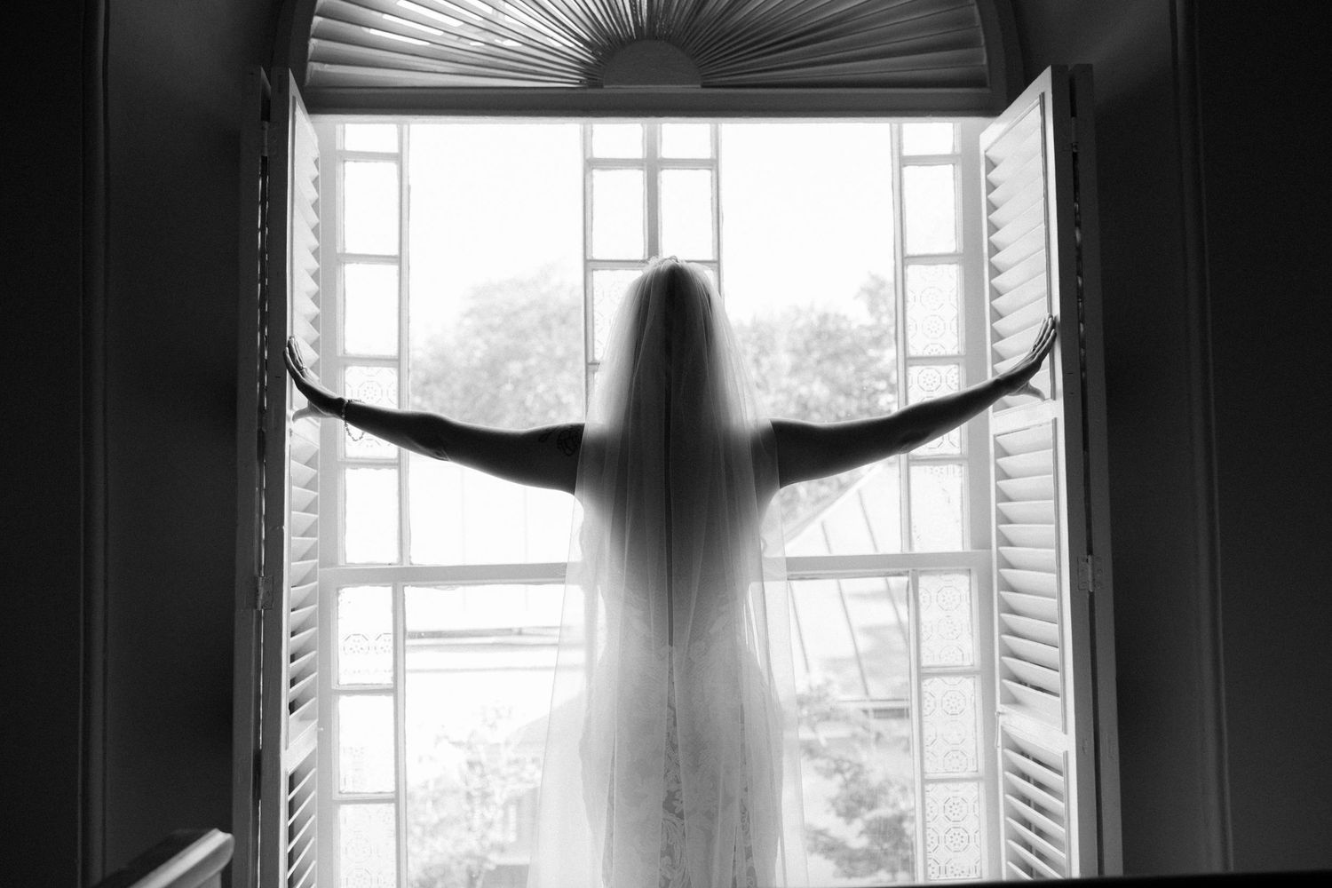Silhouette of bride in wedding veil spreading arms wide at open double window in black and white.