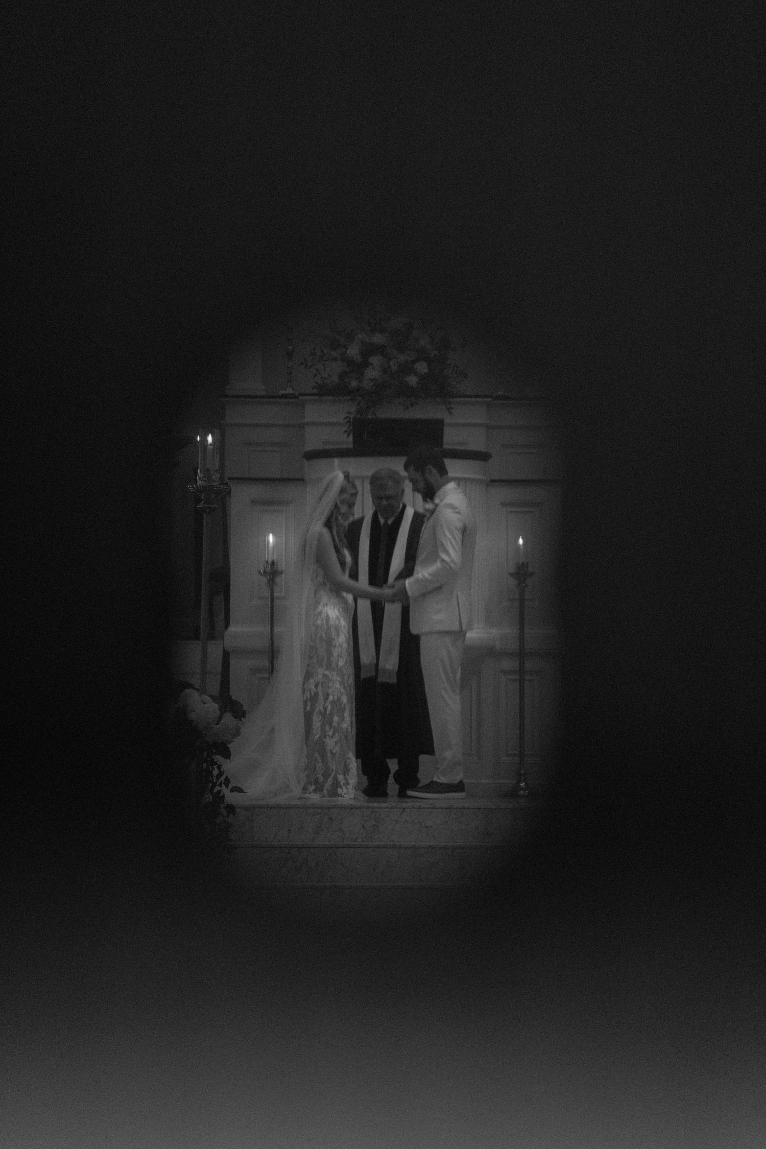 Black and white wedding ceremony photo showing bride, groom, and officiant at altar with candles in dark vignette frame.