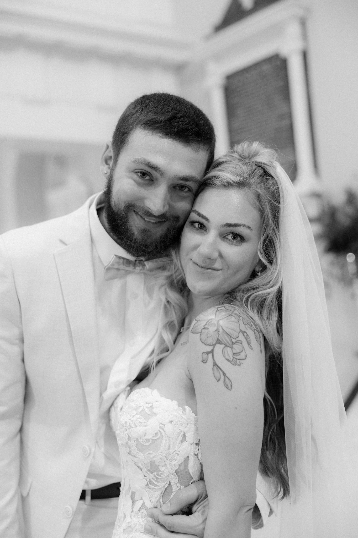 Black and white wedding portrait of smiling bride with veil and groom in white suit embracing tenderly.