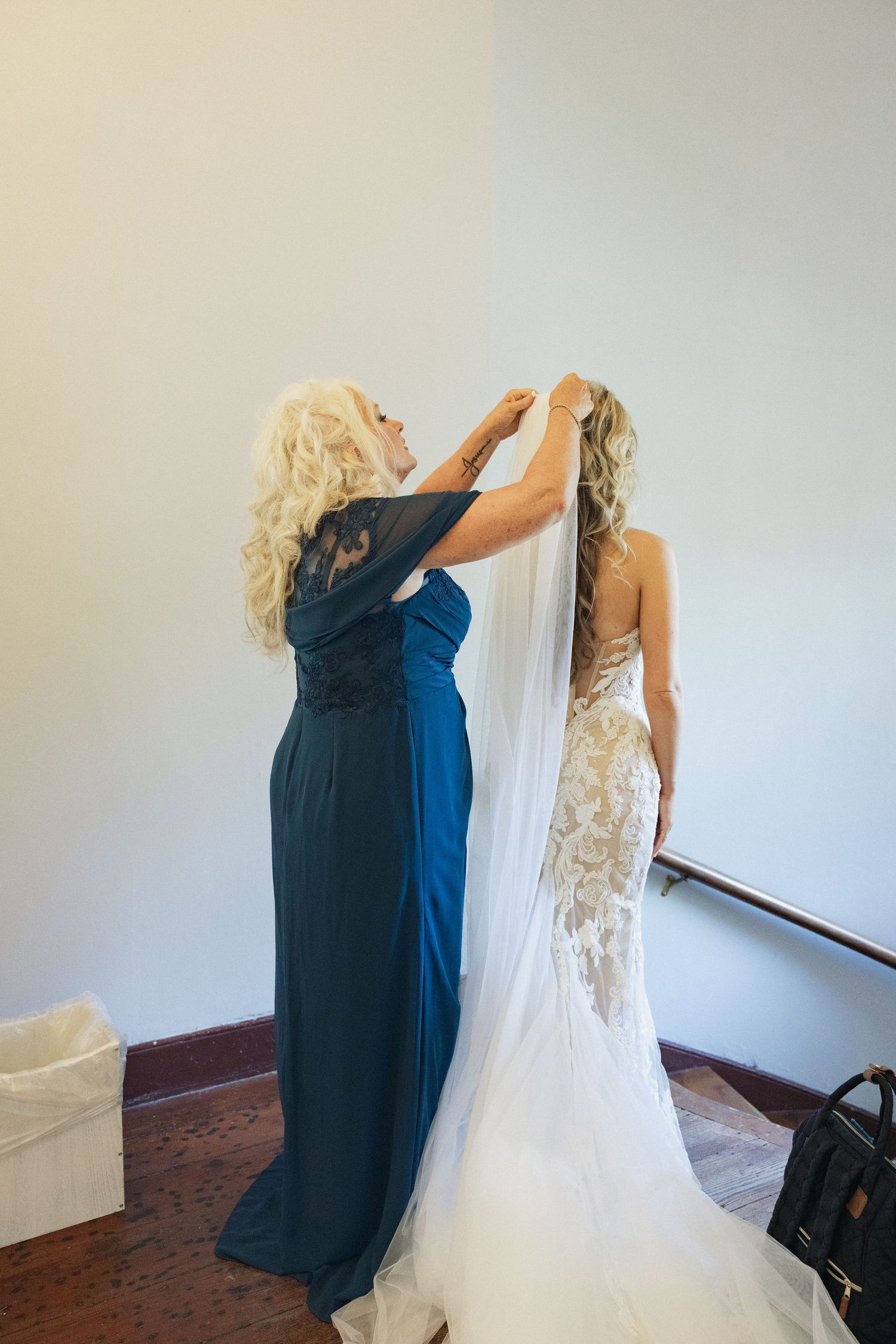 A woman in a teal gown adjusts the veil of a bride in a lace wedding dress before the ceremony.