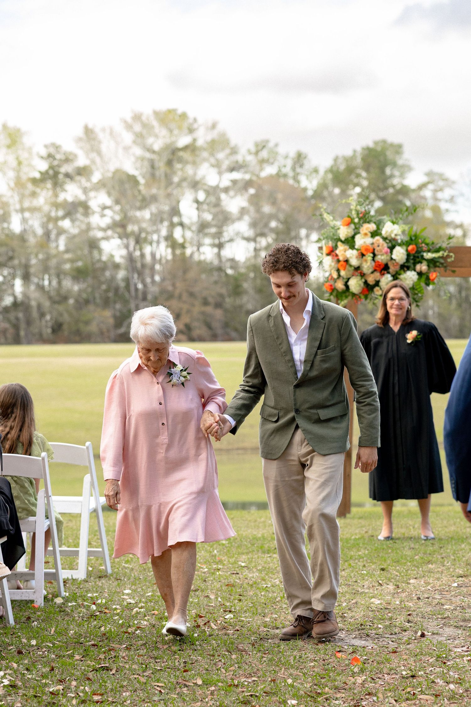 Groom escorts elderly woman in pink dress down outdoor wedding aisle with floral arch in background.