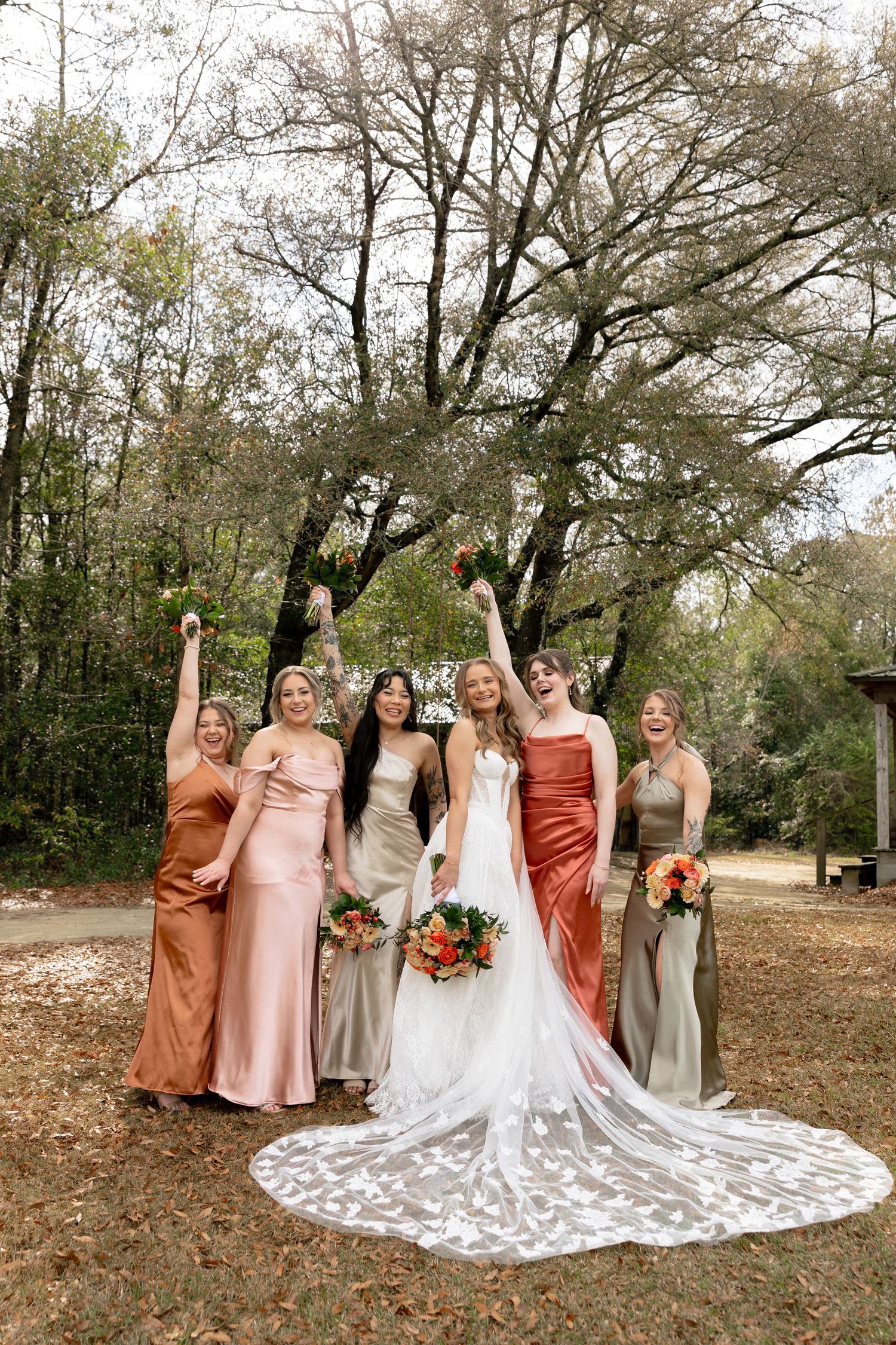 Bride and five bridesmaids celebrate joyfully with raised arms in colorful earth-tone dresses under bare trees.