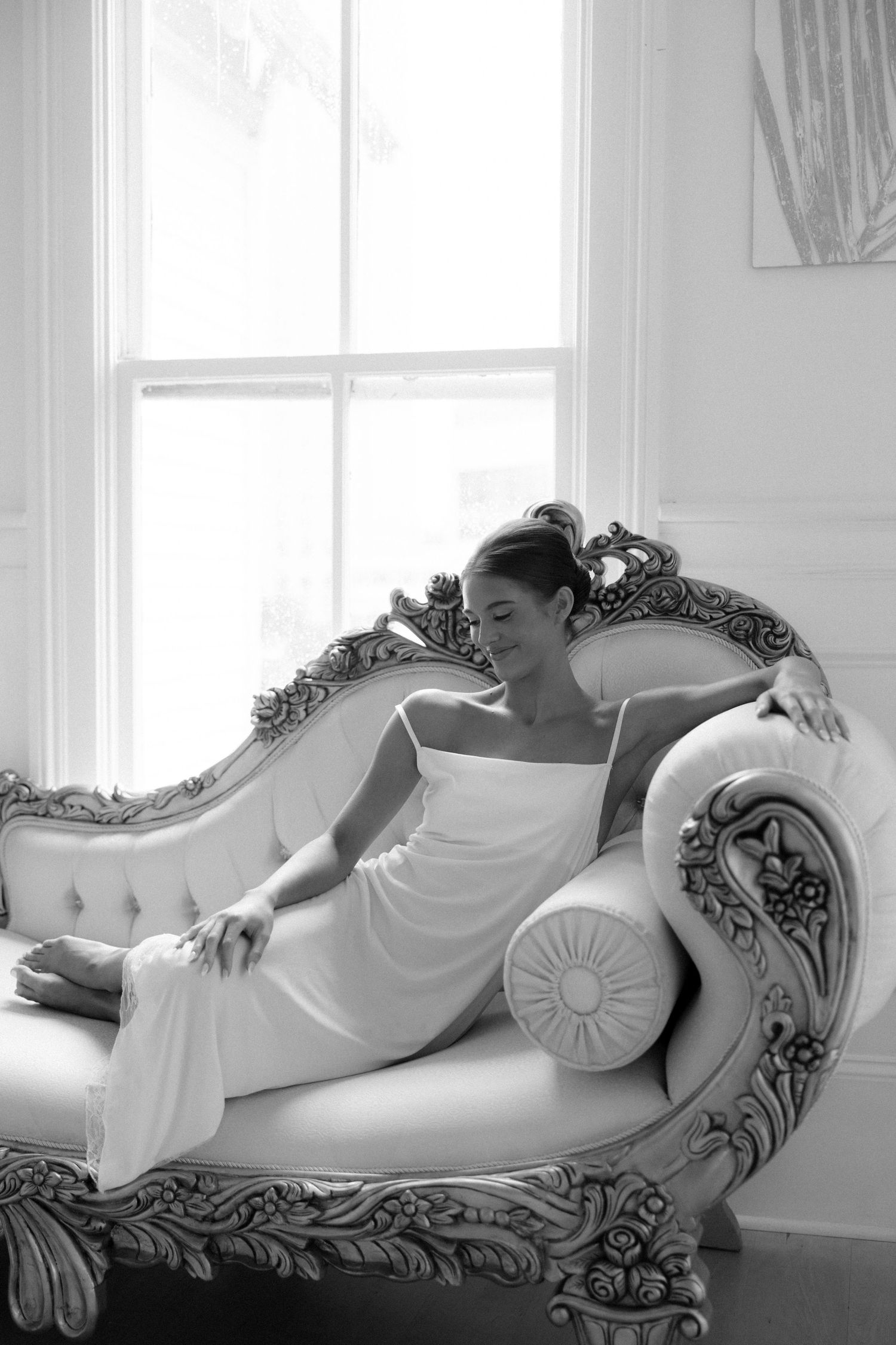 Black and white photo of a woman in a white dress reclining gracefully on an ornate vintage chaise lounge by a bright window.