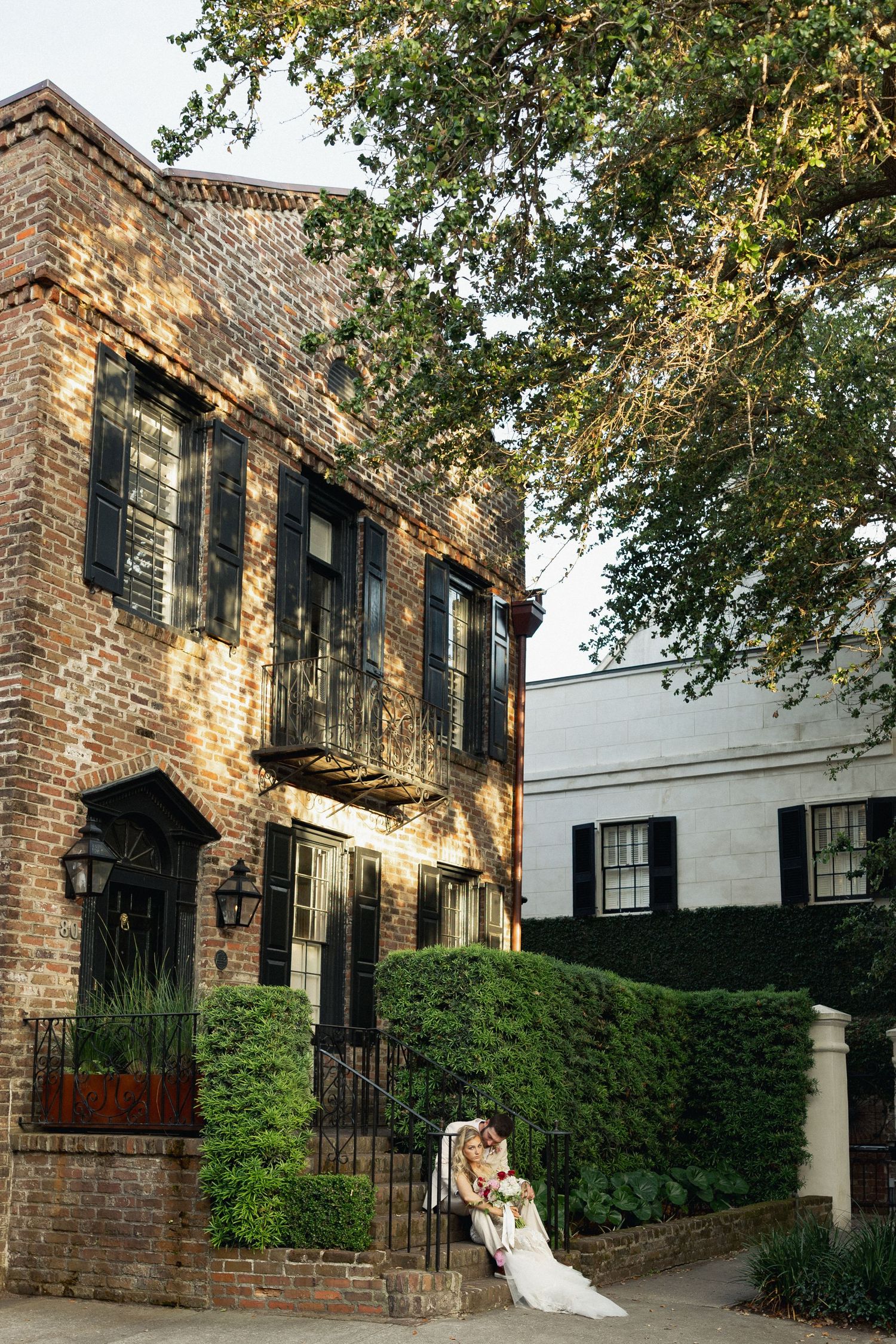Bride and groom pose outside a historic brick building with black shutters and lush greenery on their wedding day.