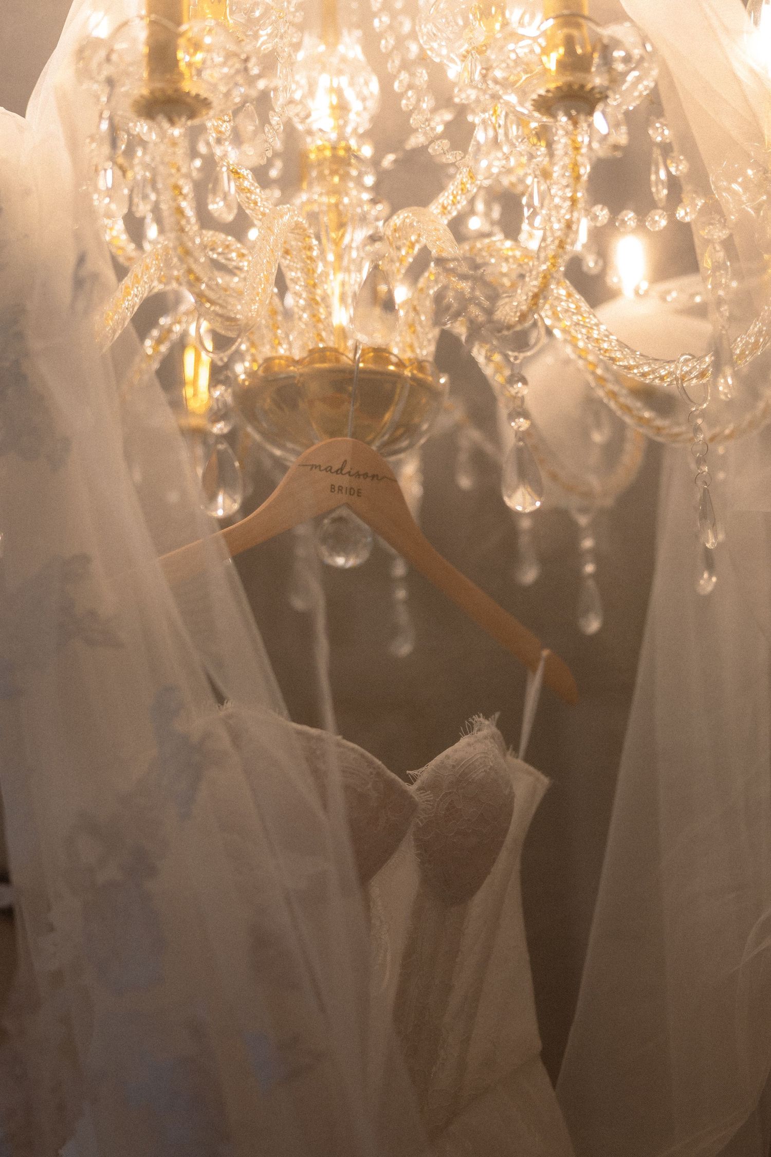 Elegant wedding dress on a wooden hanger beneath a sparkling crystal chandelier in a bridal boutique.