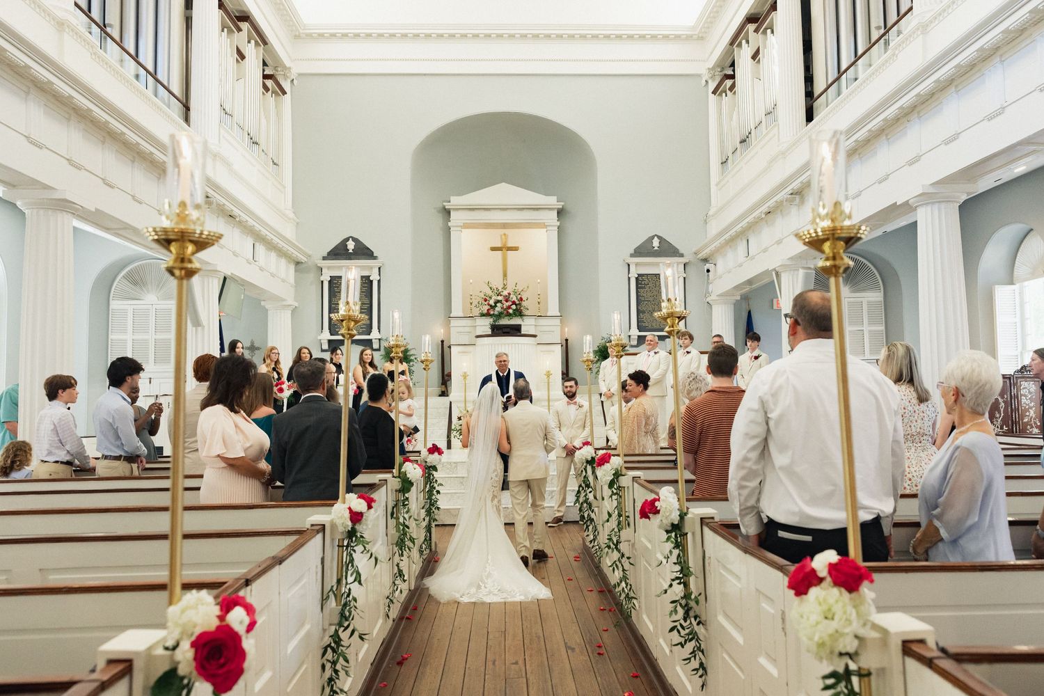 Elegant church wedding ceremony with guests seated in white pews, floral decorations, and couple at the altar.