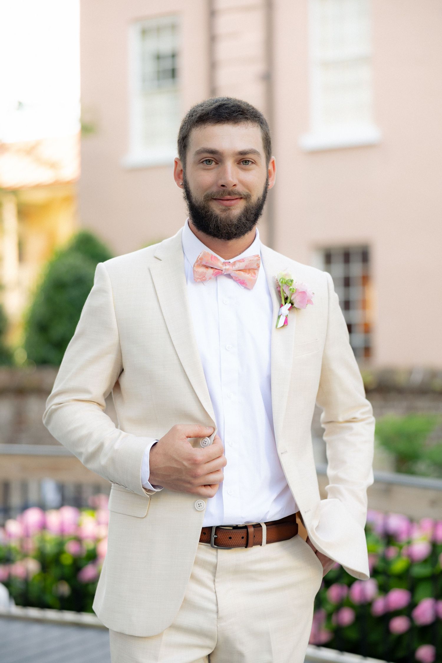Bearded groom in cream suit with pink bow tie and boutonniere poses outdoors at wedding venue.