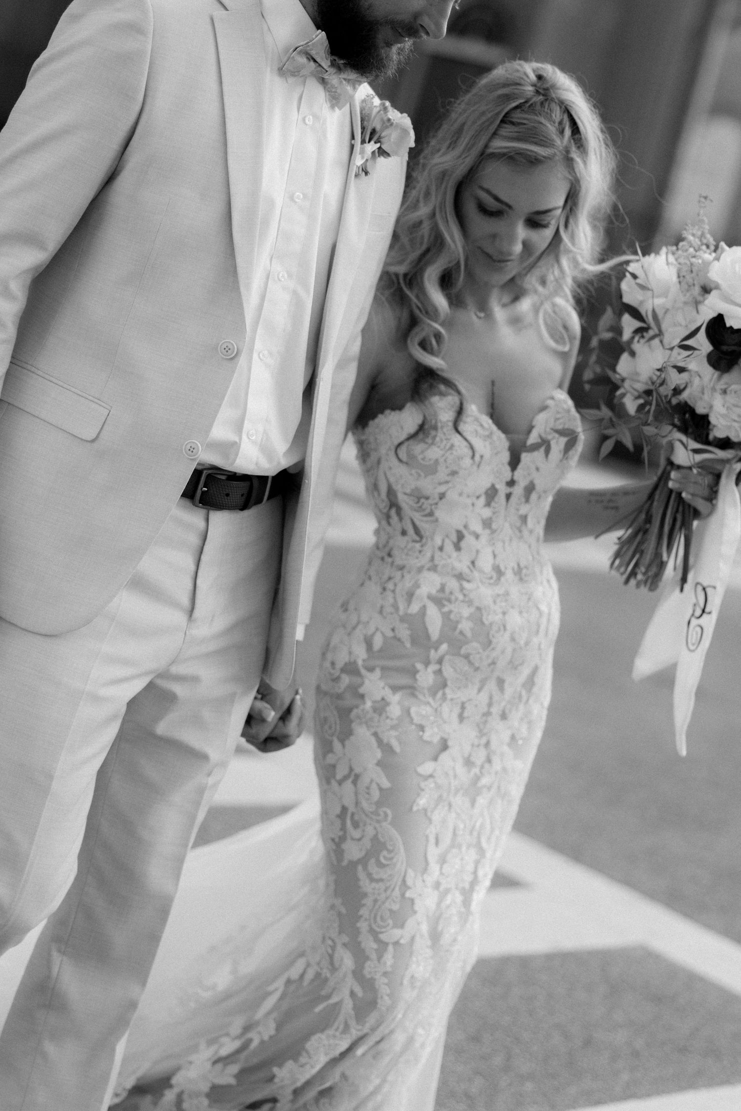 Black and white photo of bride in floral lace wedding gown holding bouquet beside groom in light suit.