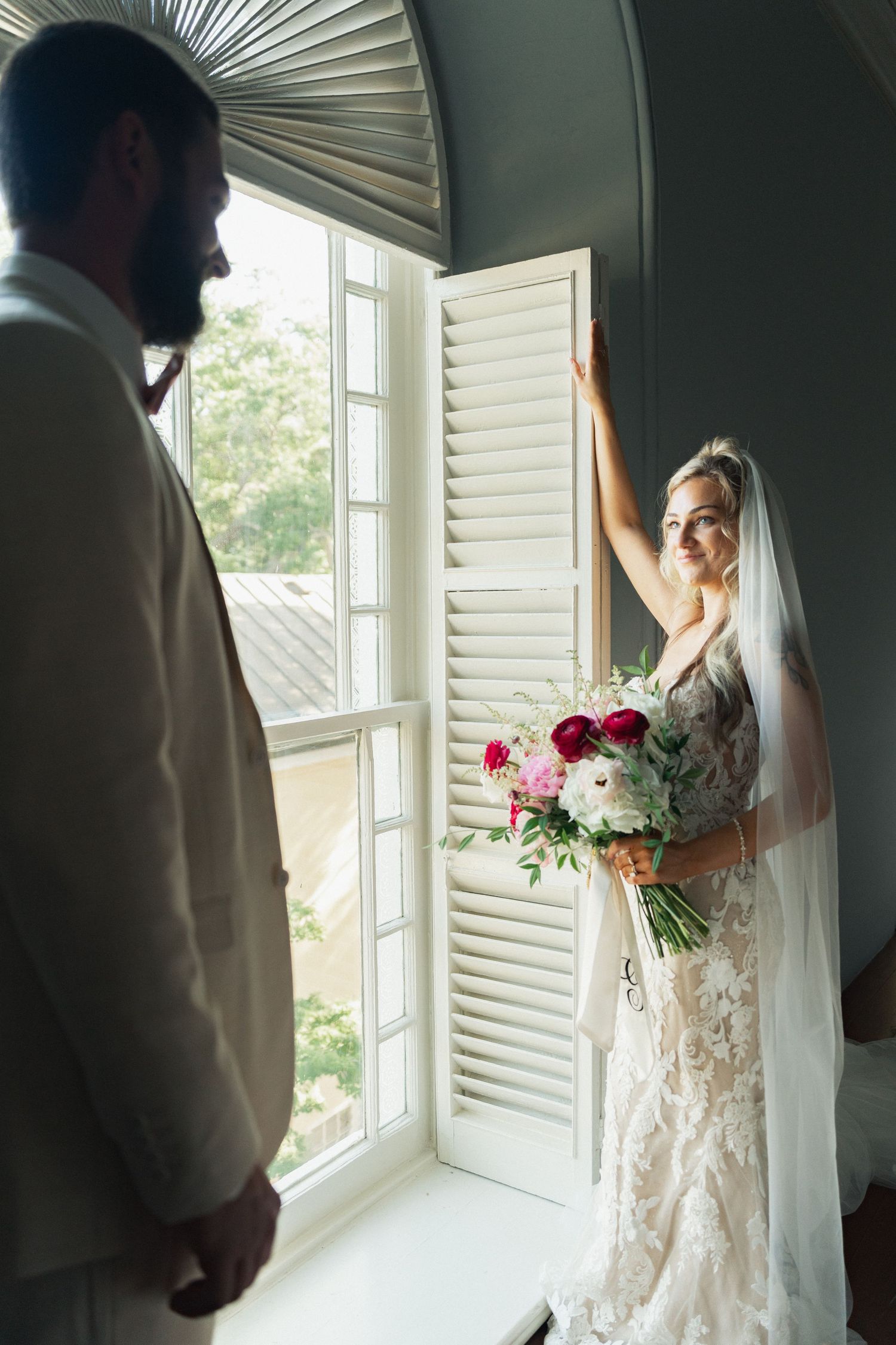 Bride in lace gown holds bouquet by window while groom in white suit looks on during wedding day first look.
