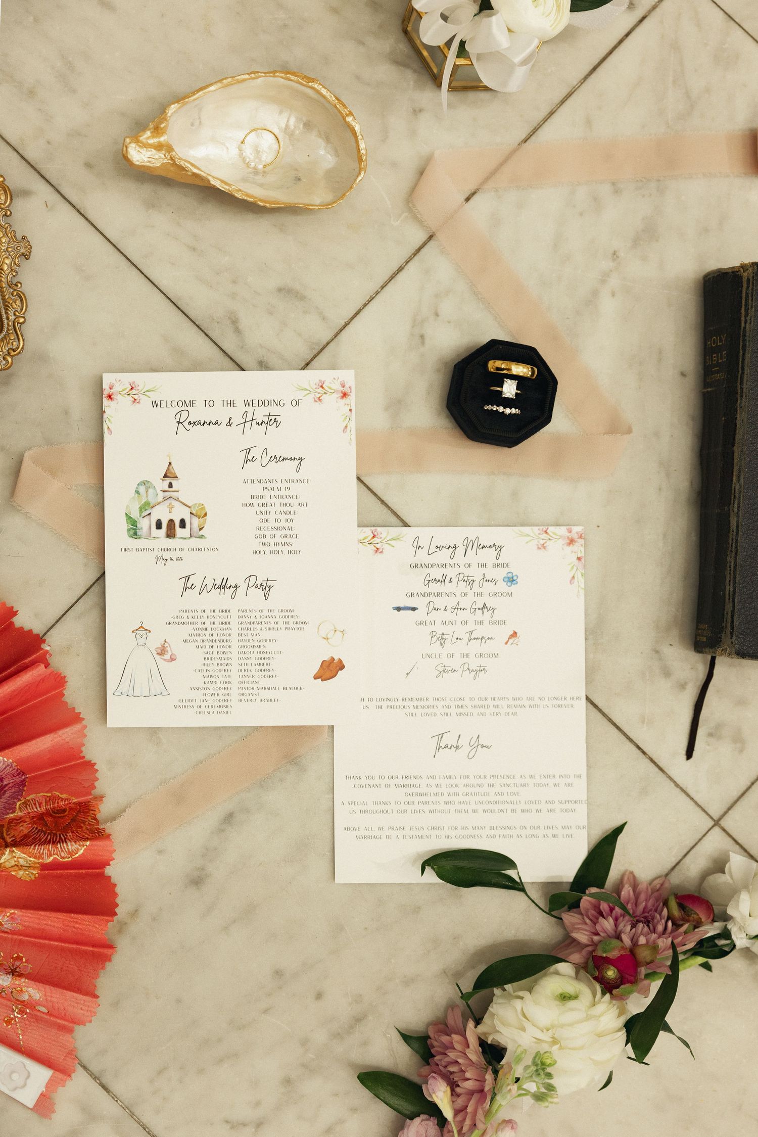 Wedding ceremony programs with illustrated church design displayed alongside red fan, rings, and flowers on marble.