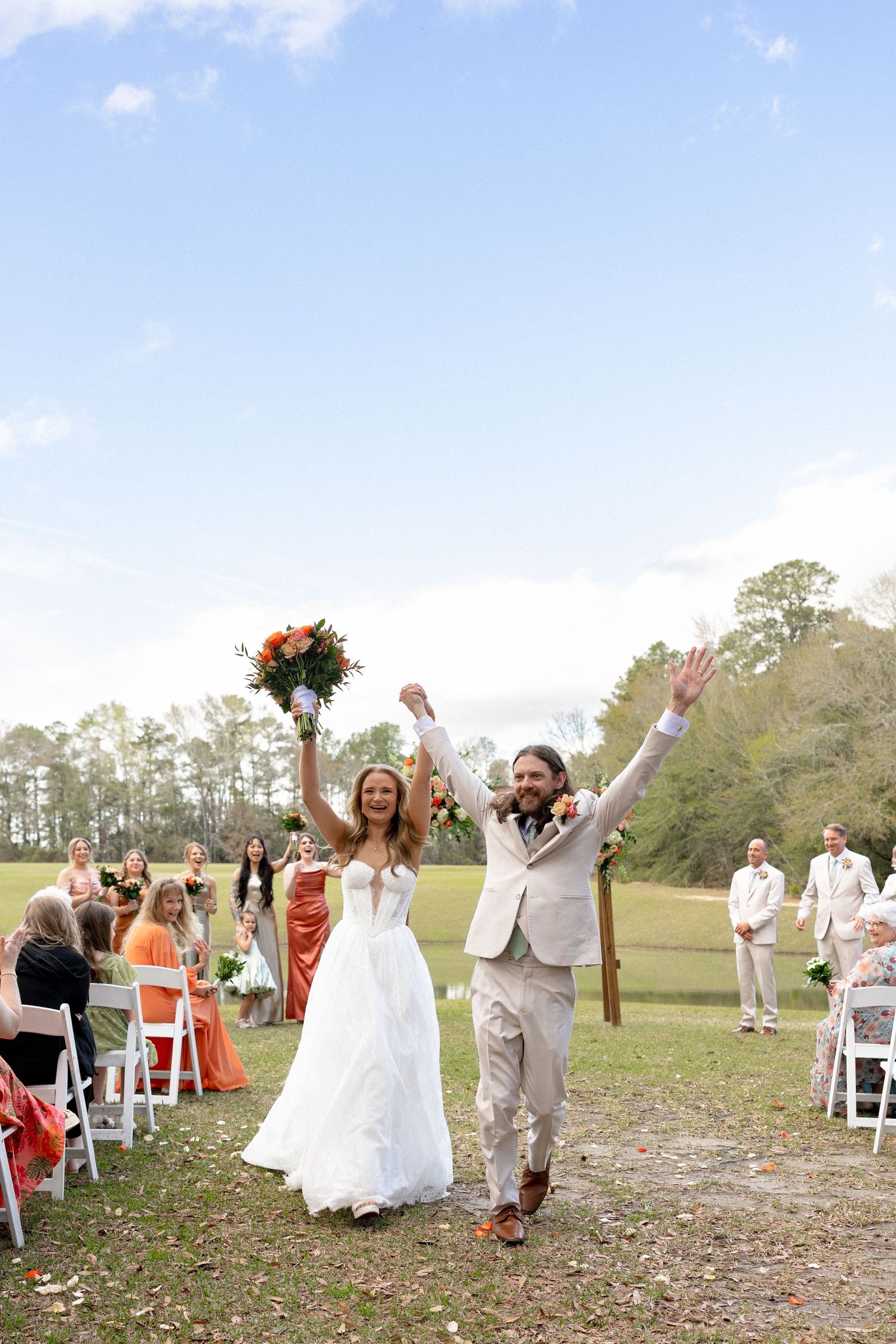 Jubilant newlyweds raise hands in celebration while walking down outdoor aisle with wedding guests seated nearby.