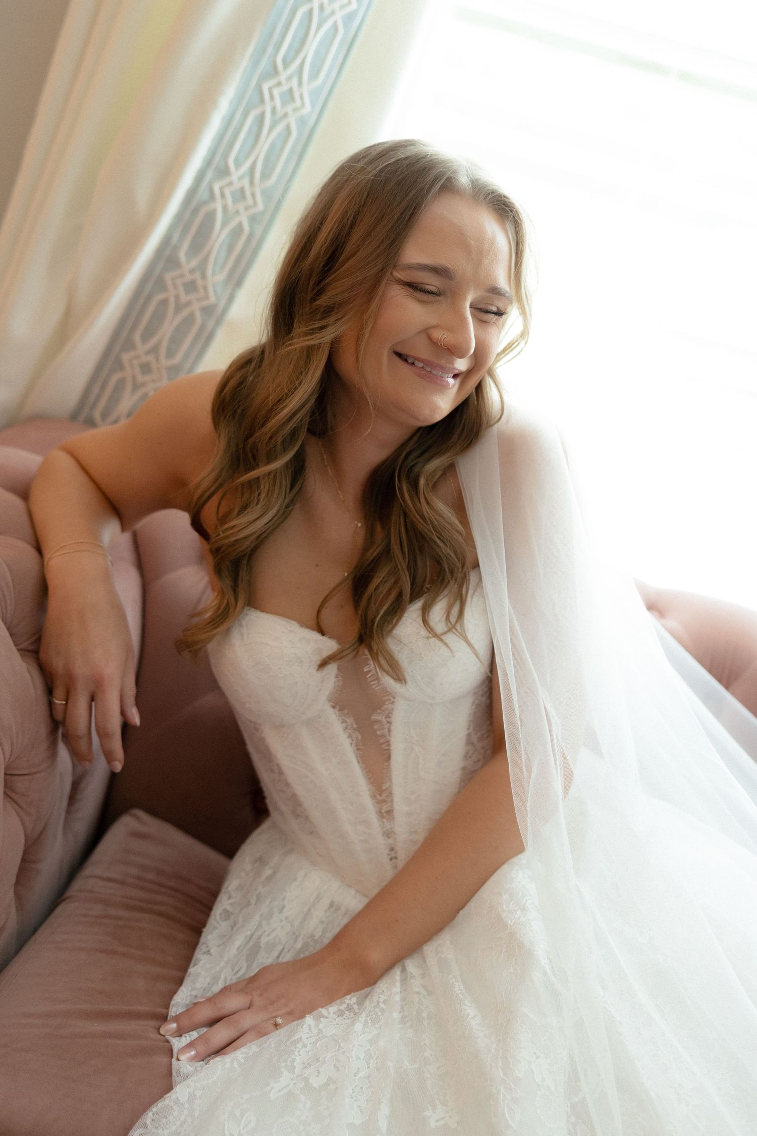 Smiling bride in strapless lace wedding gown and veil seated on pink velvet chair by window.
