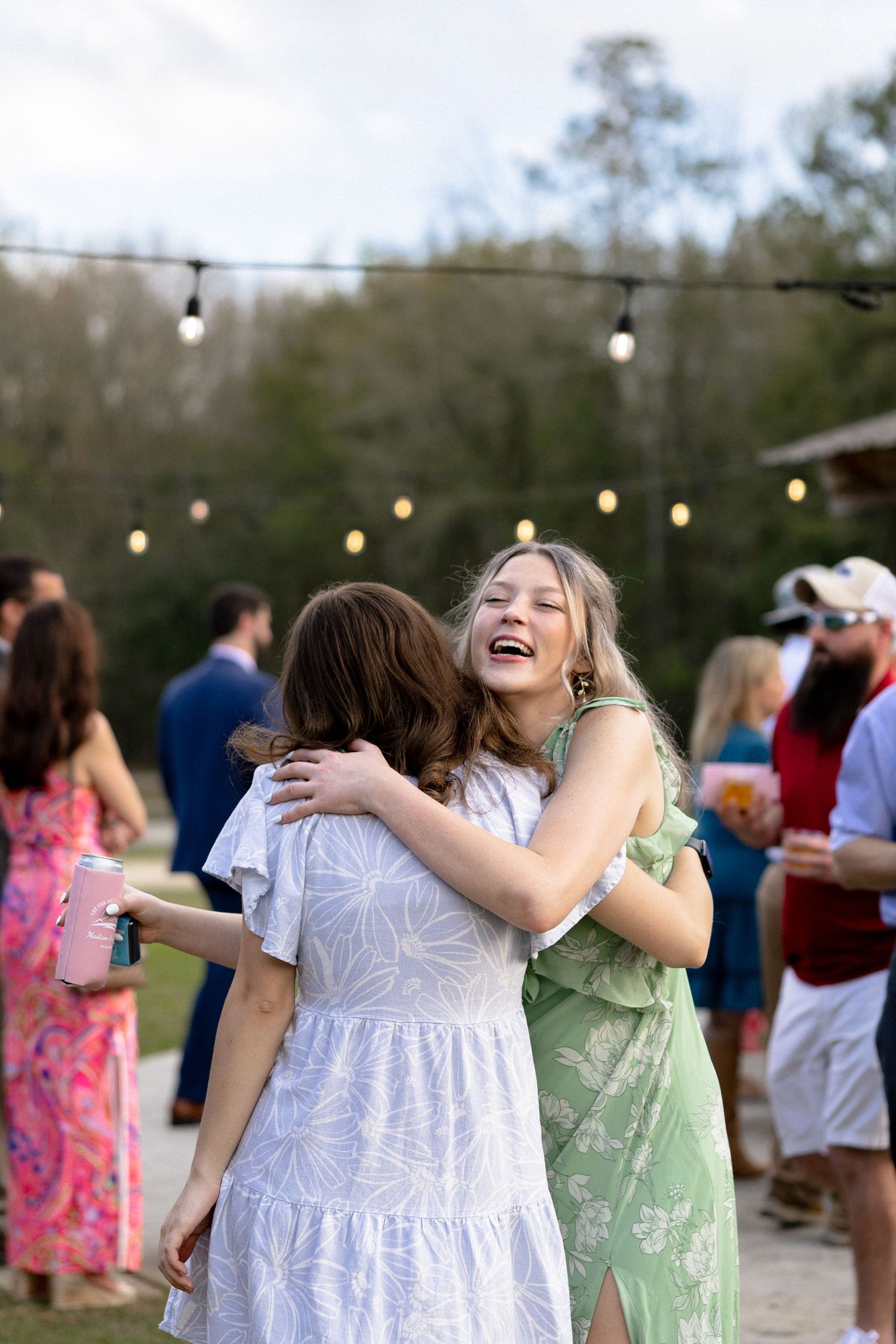 Two young women laughing and hugging at an outdoor wedding reception with string lights.