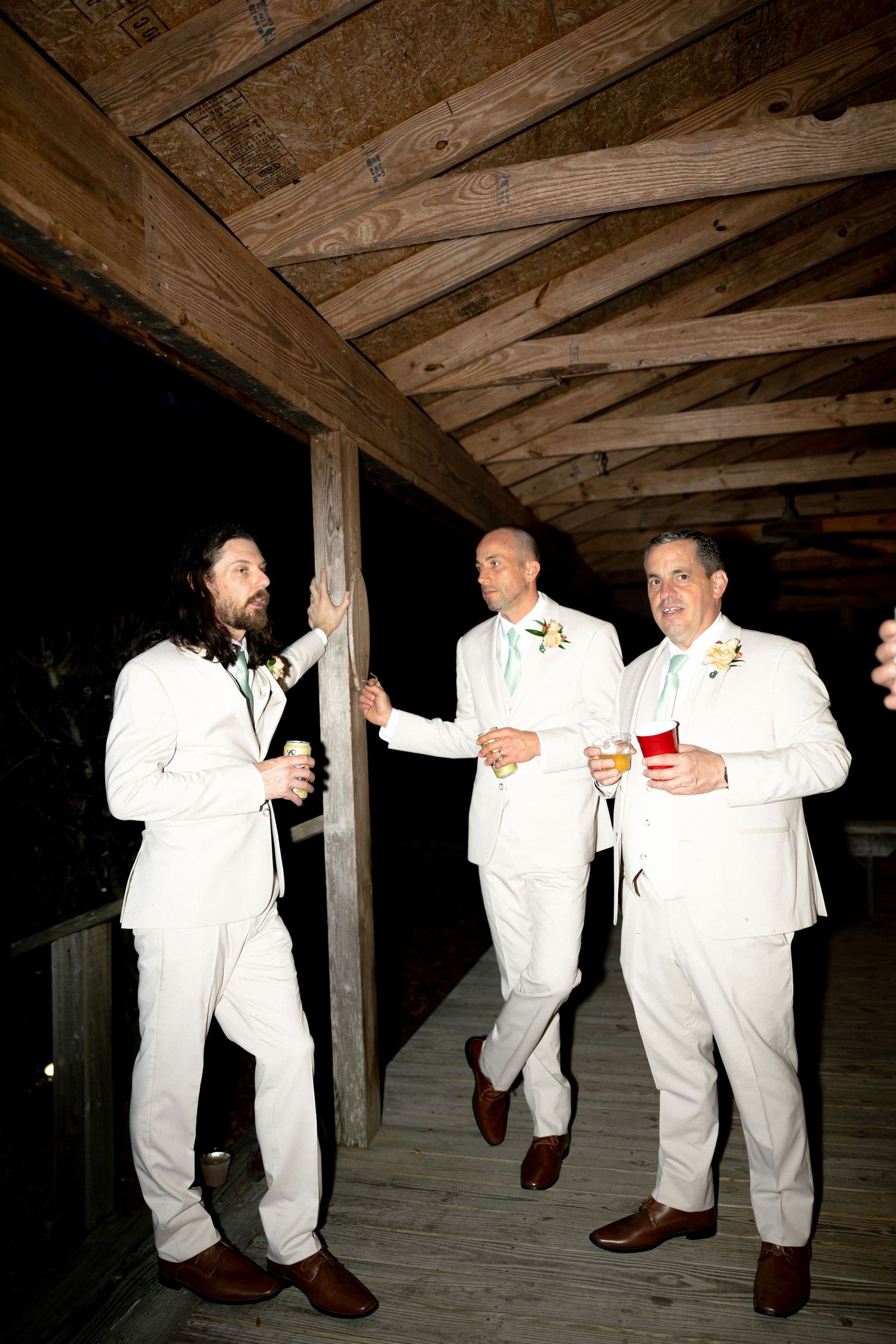 Groom and groomsmen in white suits laugh and celebrate together on a wooden deck at night.