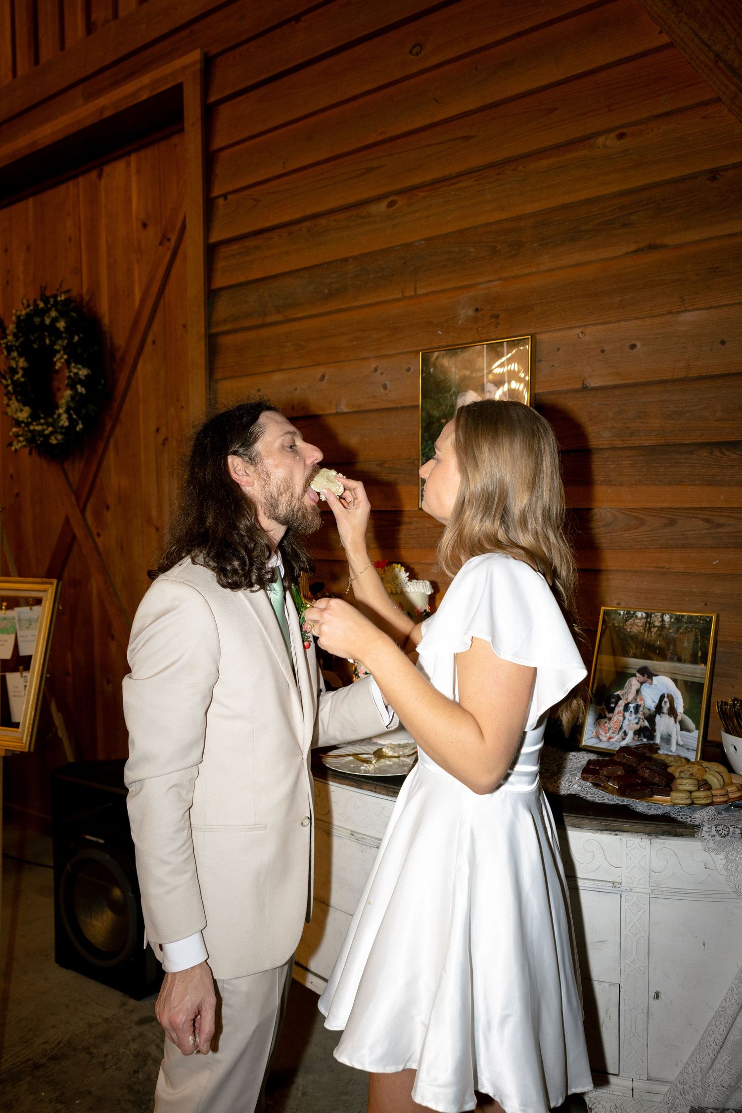 Bride in white dress feeds wedding cake to groom in beige suit at rustic barn wedding reception.
