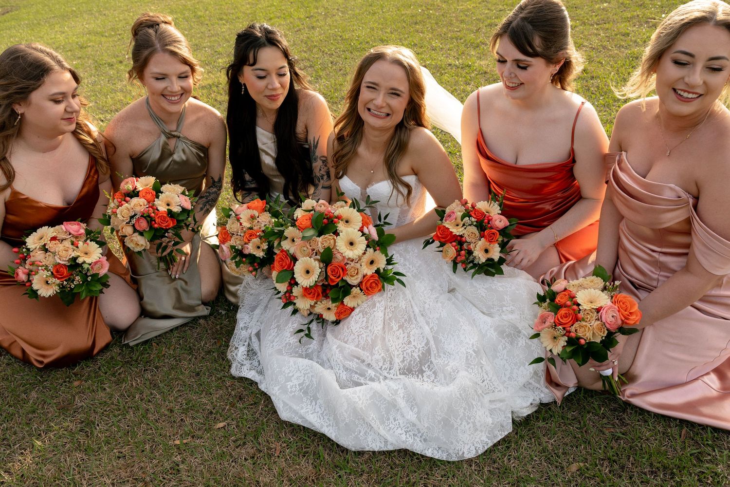 Bride in white lace gown sitting on grass with bridesmaids holding peach and orange bouquets smiling.