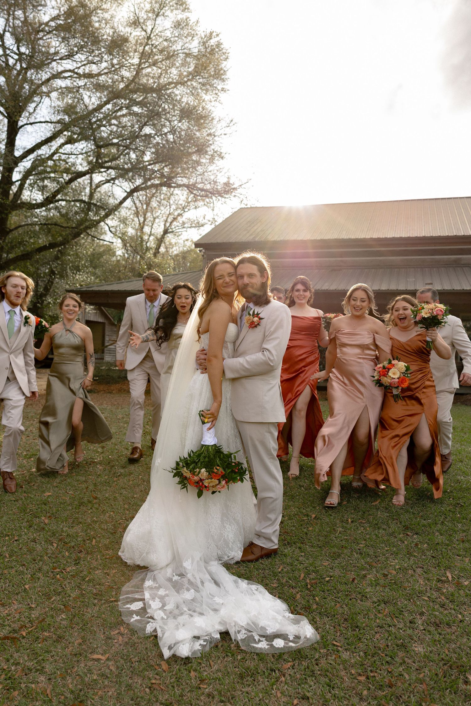 Groom embraces bride as excited wedding party runs toward camera in golden sunset light near rustic barn.