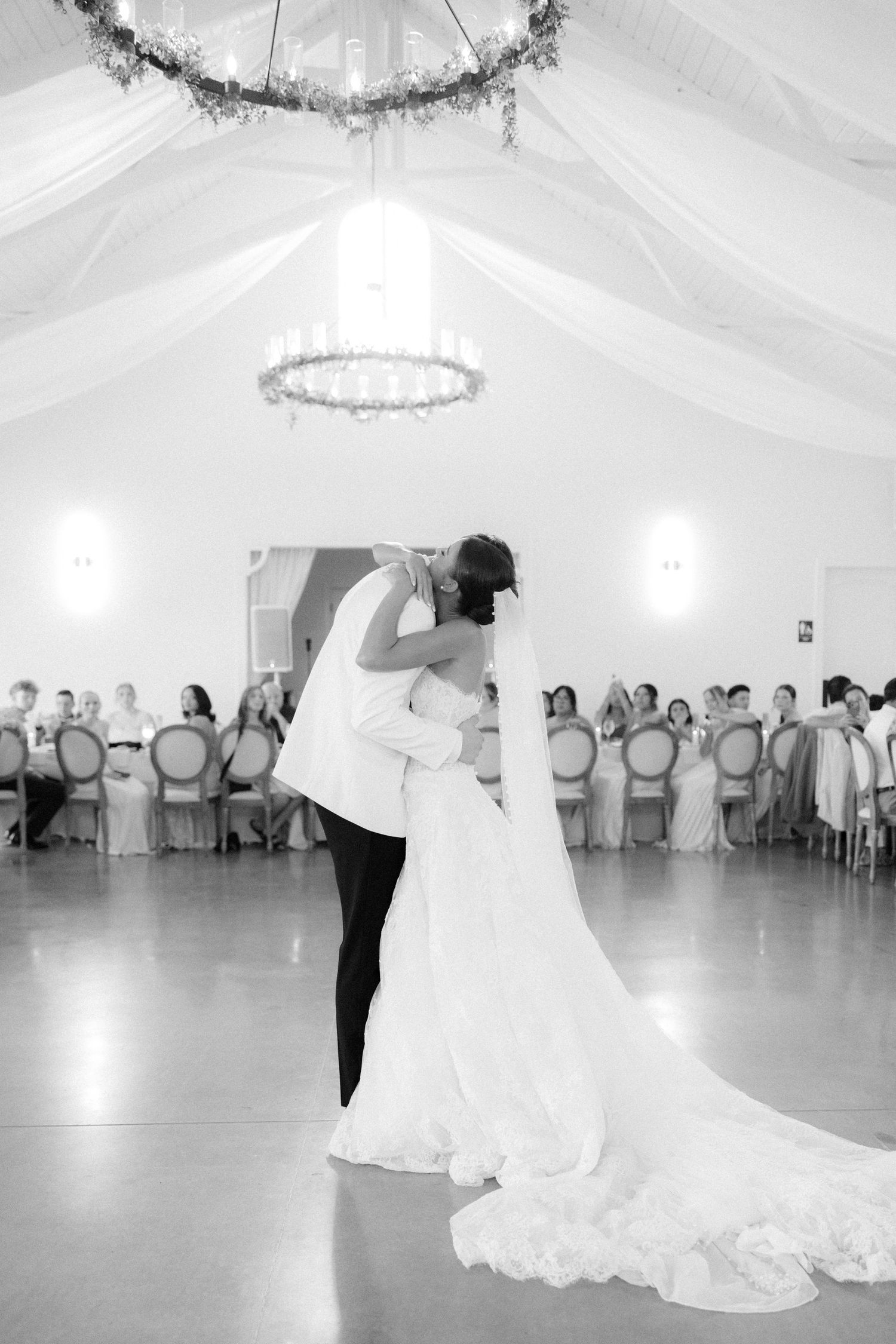 Black and white photo of bride and groom sharing romantic first dance embrace before wedding guests.