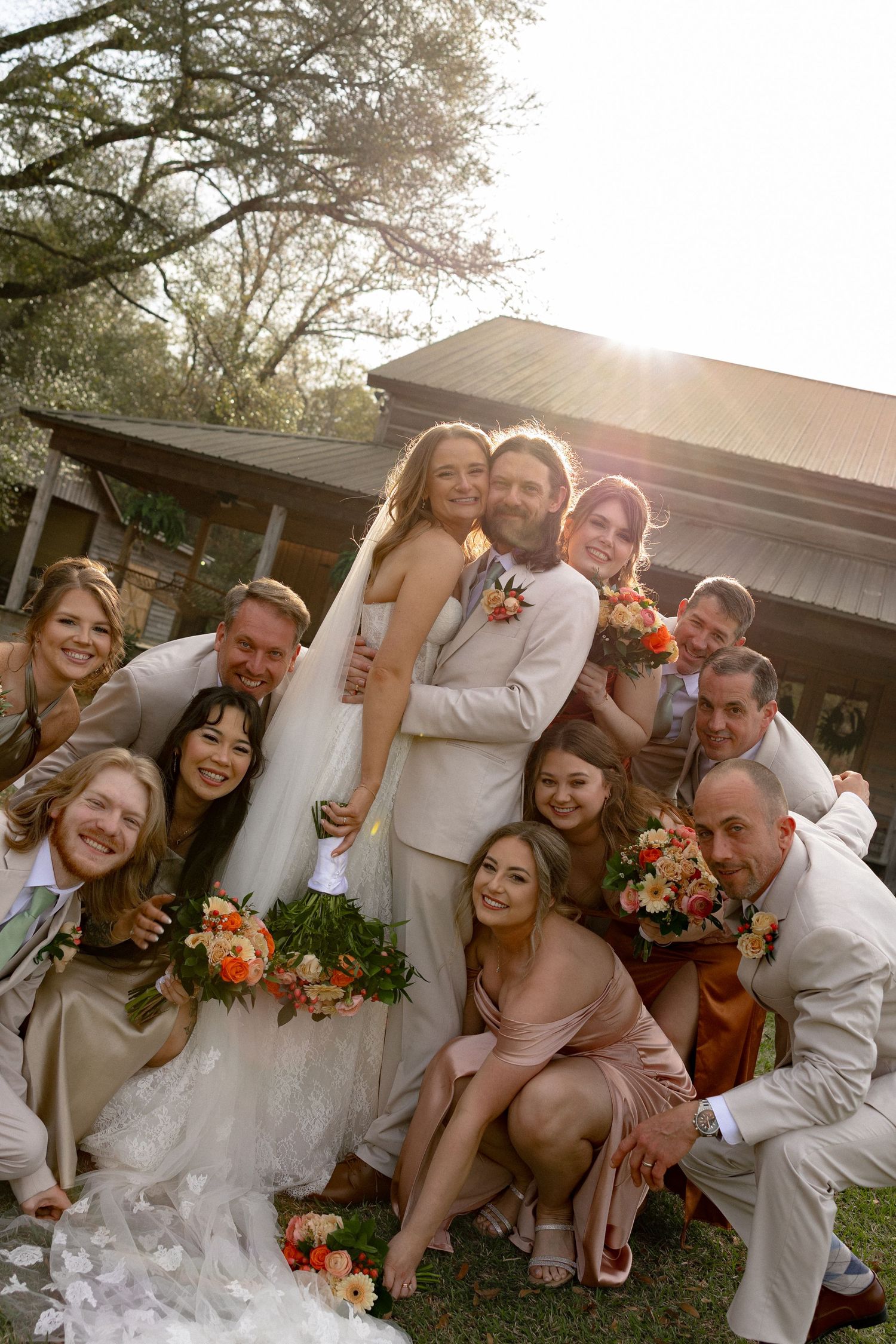 Joyful wedding party group photo with bride and groom surrounded by bridesmaids and groomsmen in golden sunlight.